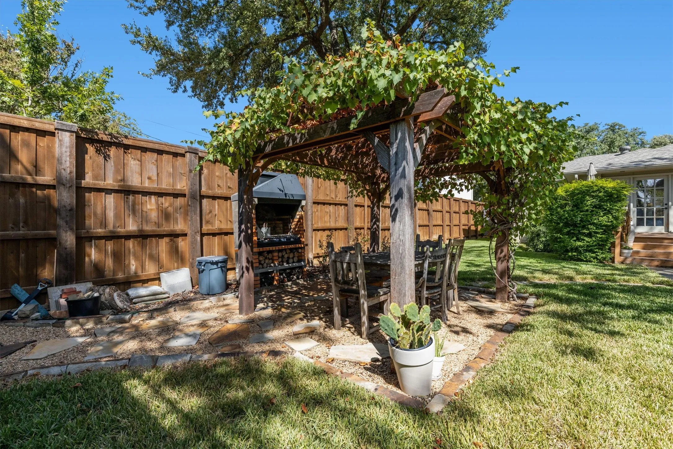 Pergola with mature vine coverage, stone and gravel patio, wood privacy fencing, and a manicured lawn