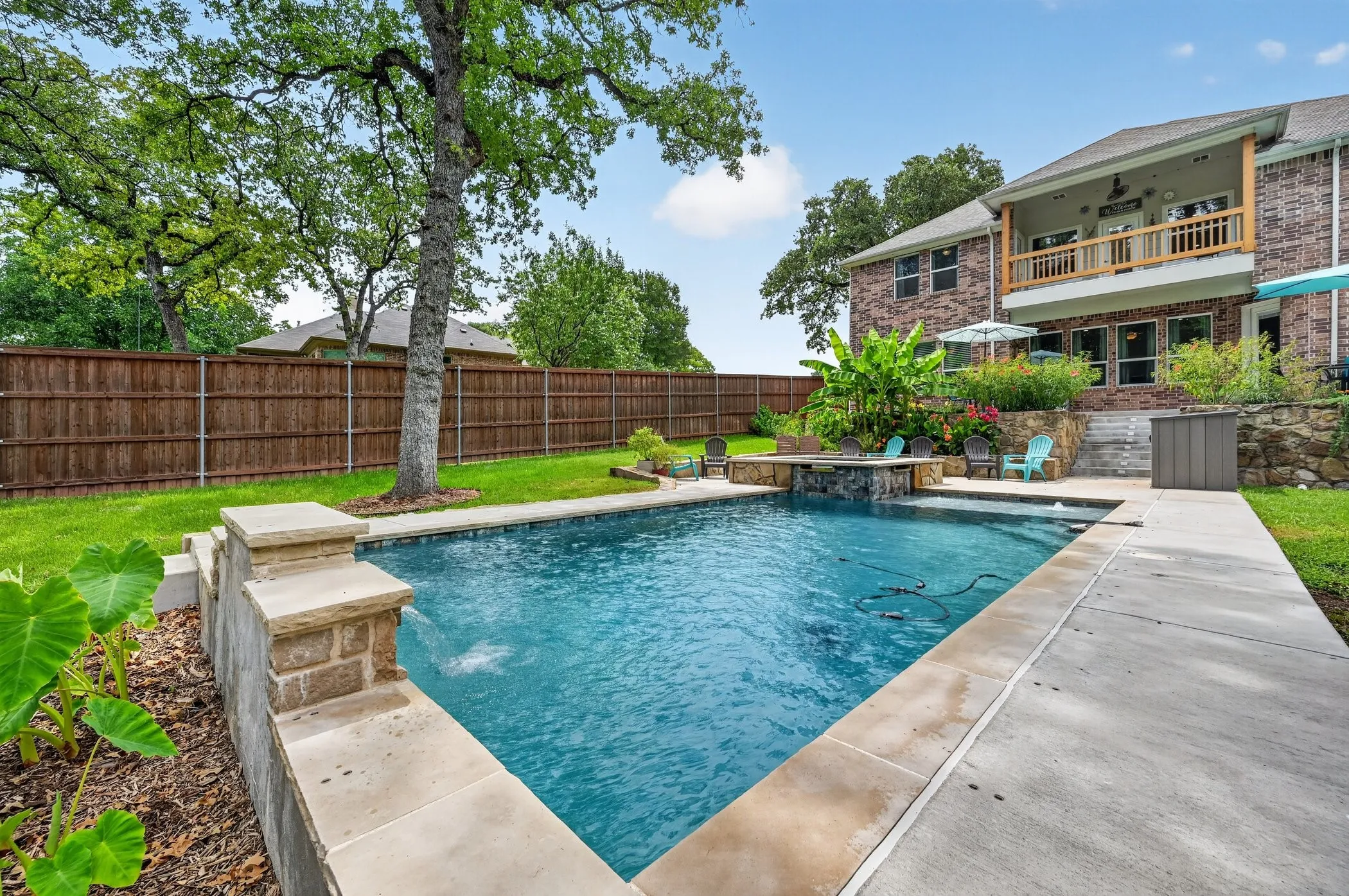 Expansive backyard featuring a custom-shaped pool, stone-clad retaining walls, a wood privacy fence, mature trees, and a two-story brick exterior with an upper-level balcony