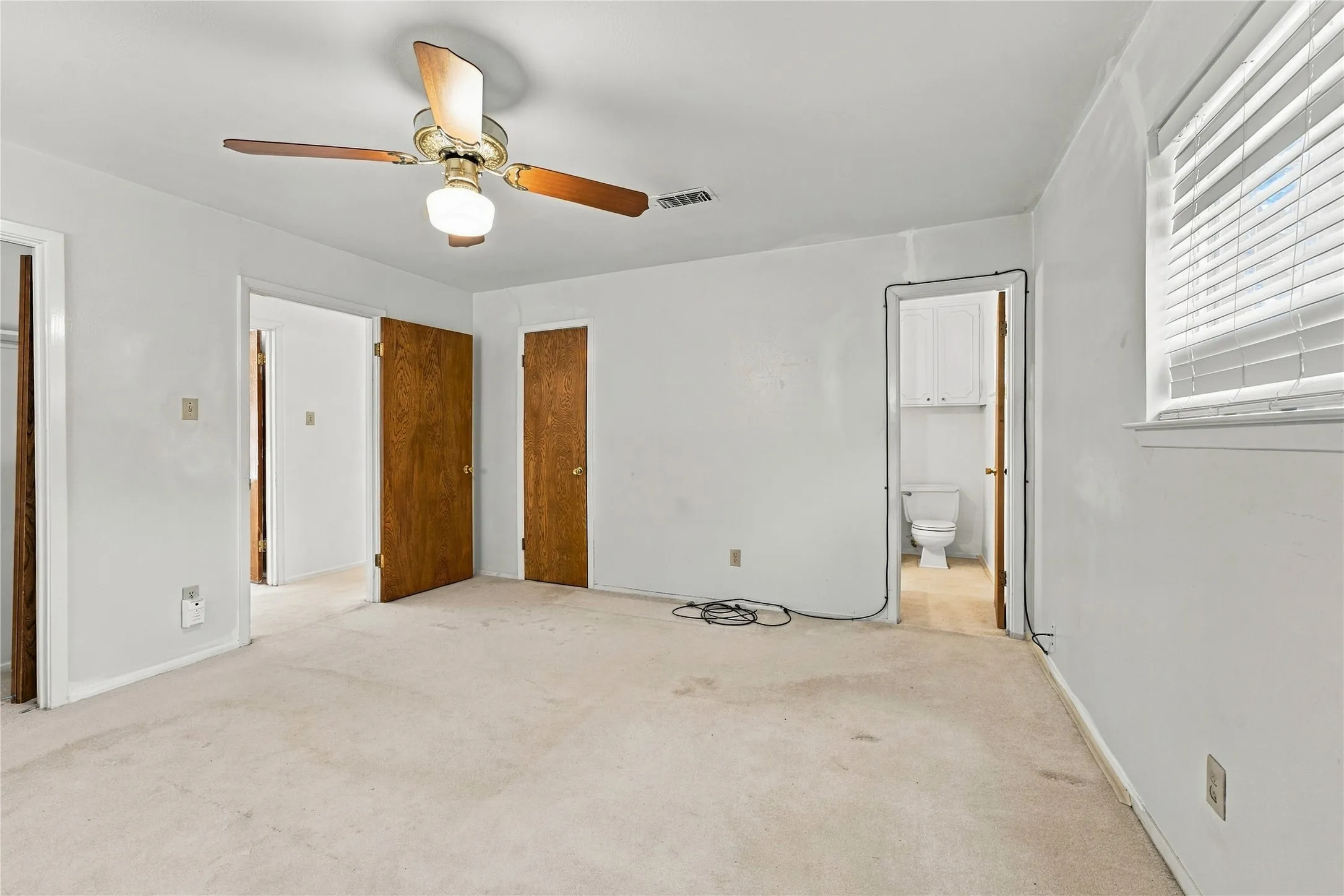 Neutral wall tones, wood-finish interior doors, ceiling fan with integrated lighting, window with blinds, and an adjacent bathroom with built-in cabinetry