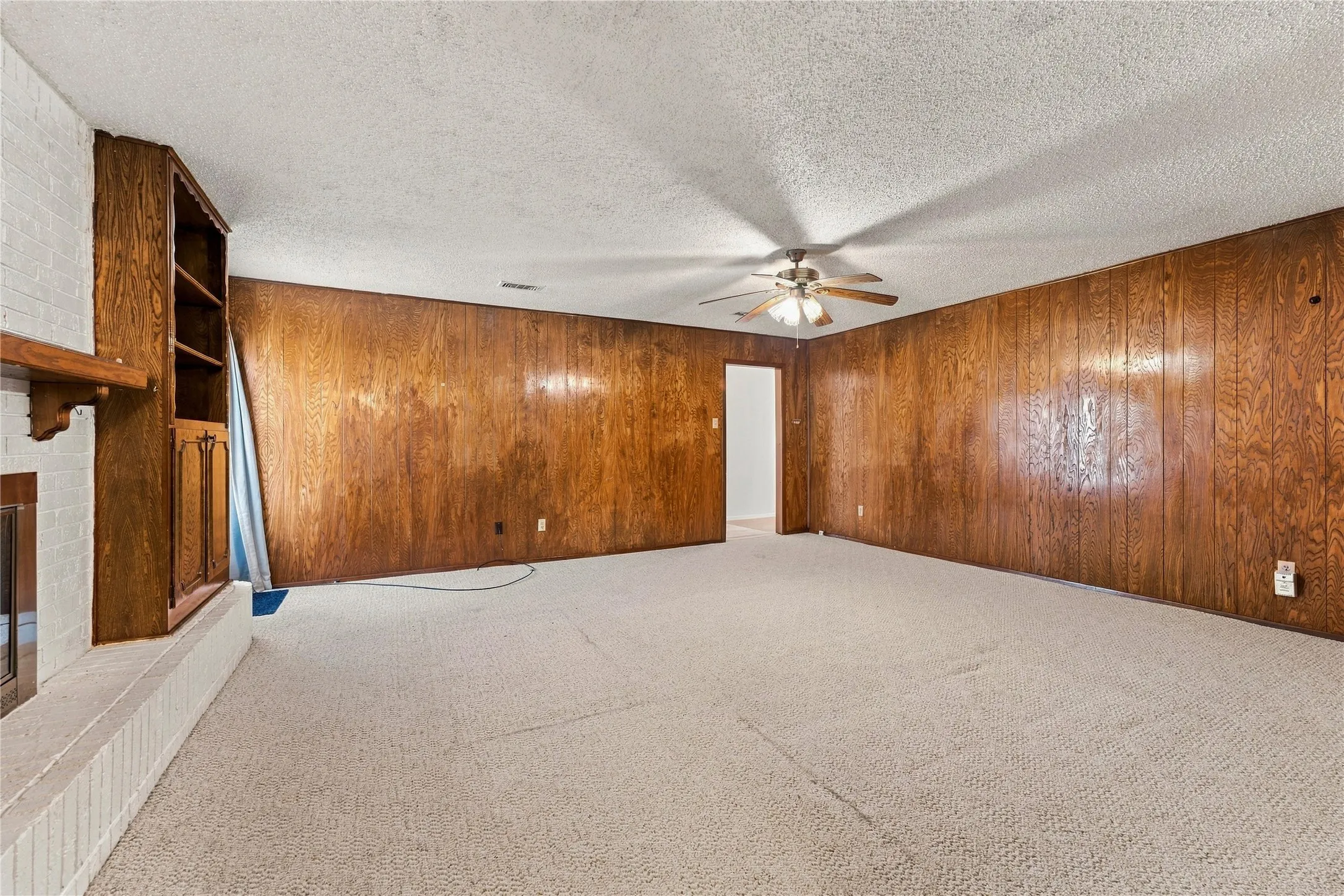 Wall paneling, built-in shelving, white brick fireplace, ceiling fan, and carpet flooring