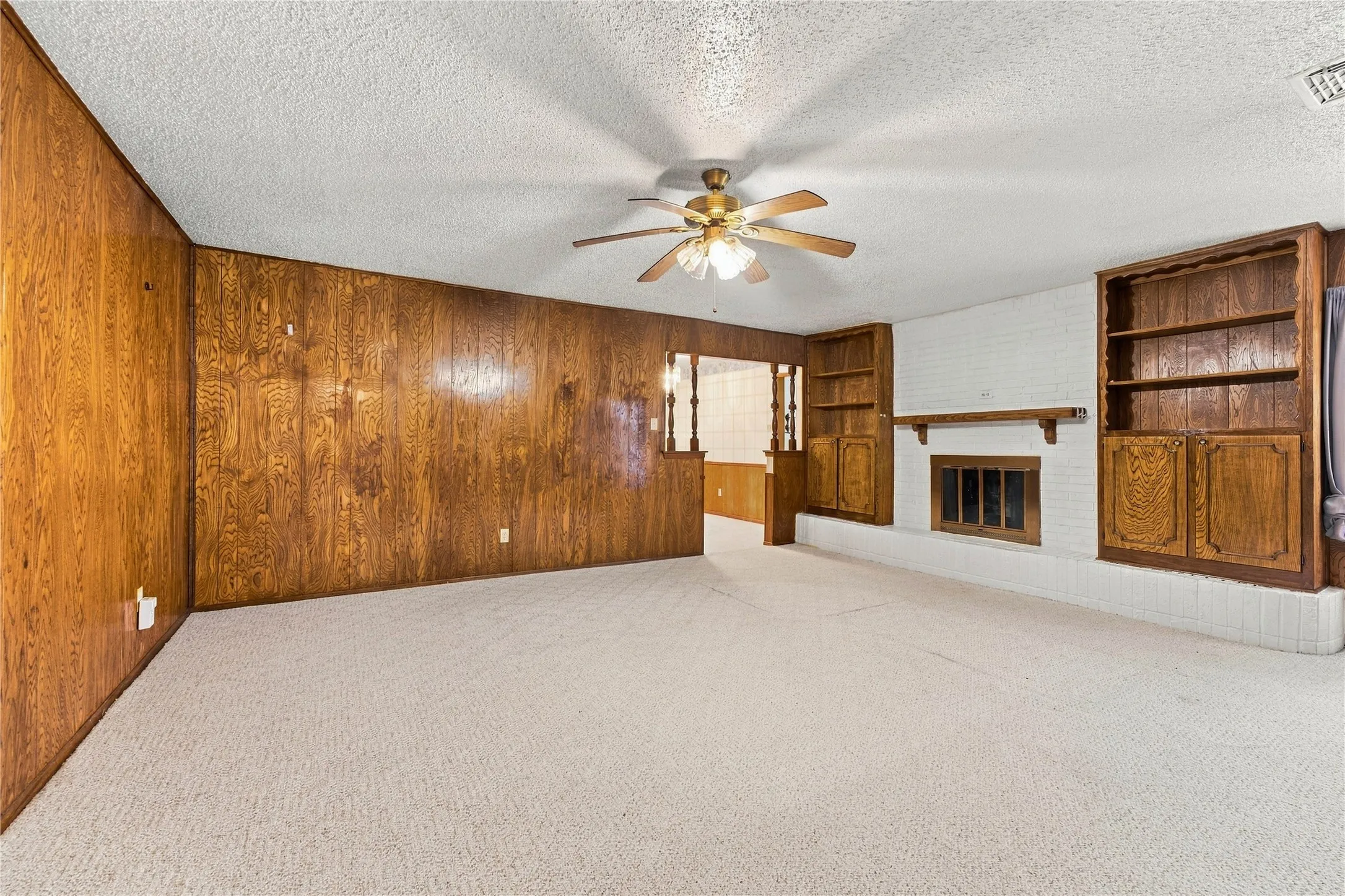 Spacious interior featuring wood paneling, an integrated fireplace with a white brick surround, built-in shelving, and a ceiling fan