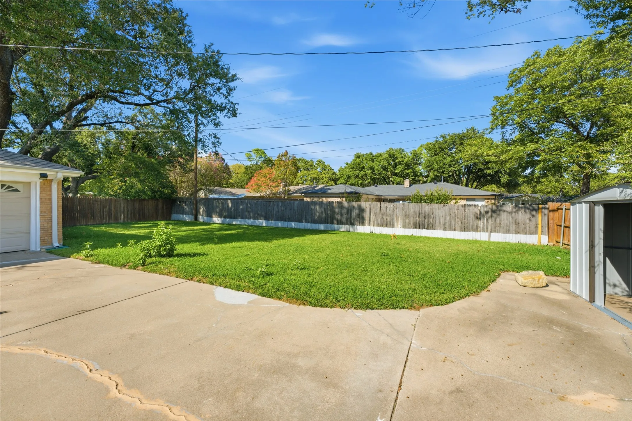 Expansive concrete patio and driveway, fenced backyard with natural grass, mature trees, and a detached storage shed