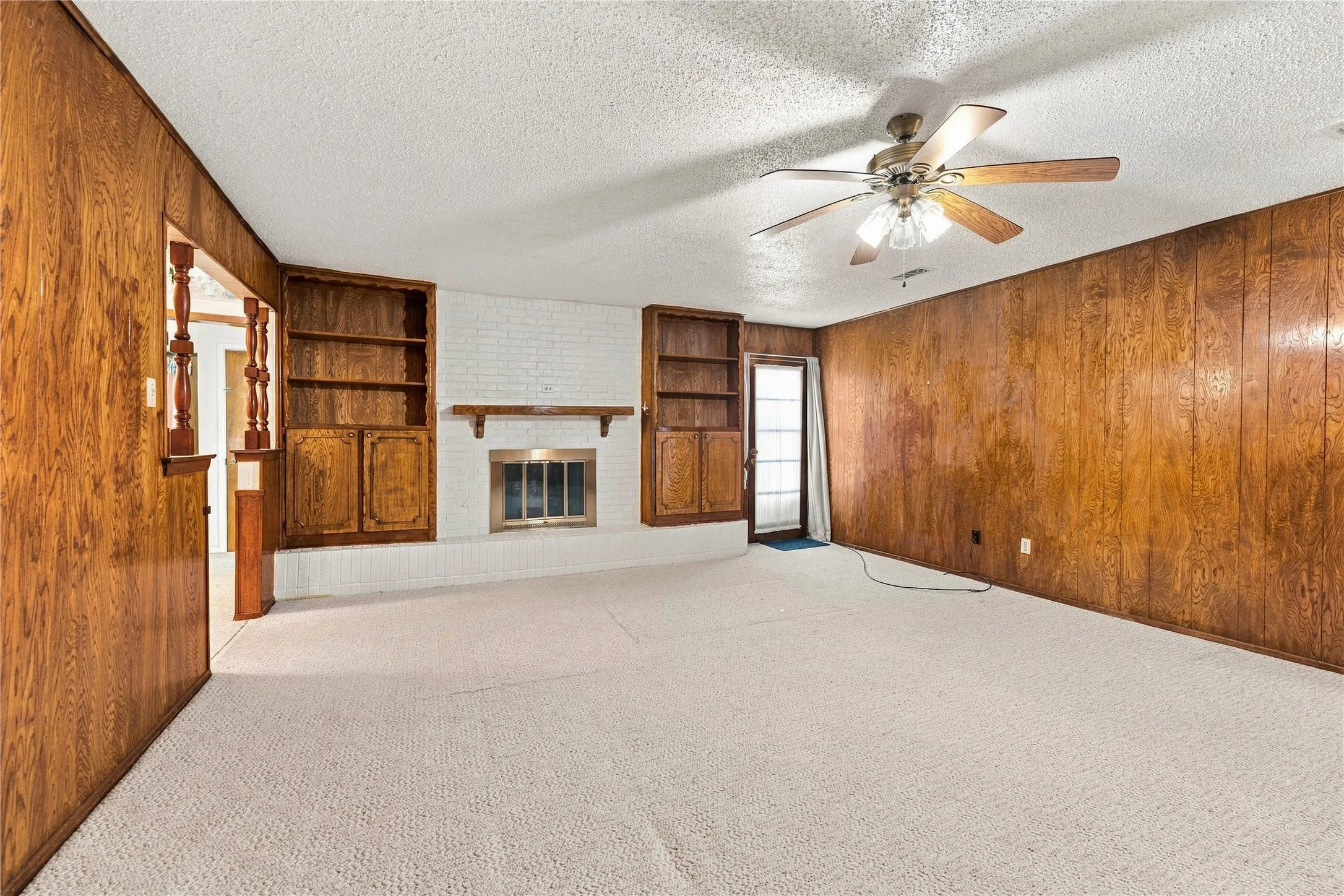 Wood-paneled walls, white brick fireplace with mantel, built-in shelving and cabinetry, ceiling fan with light fixture, and plush carpet