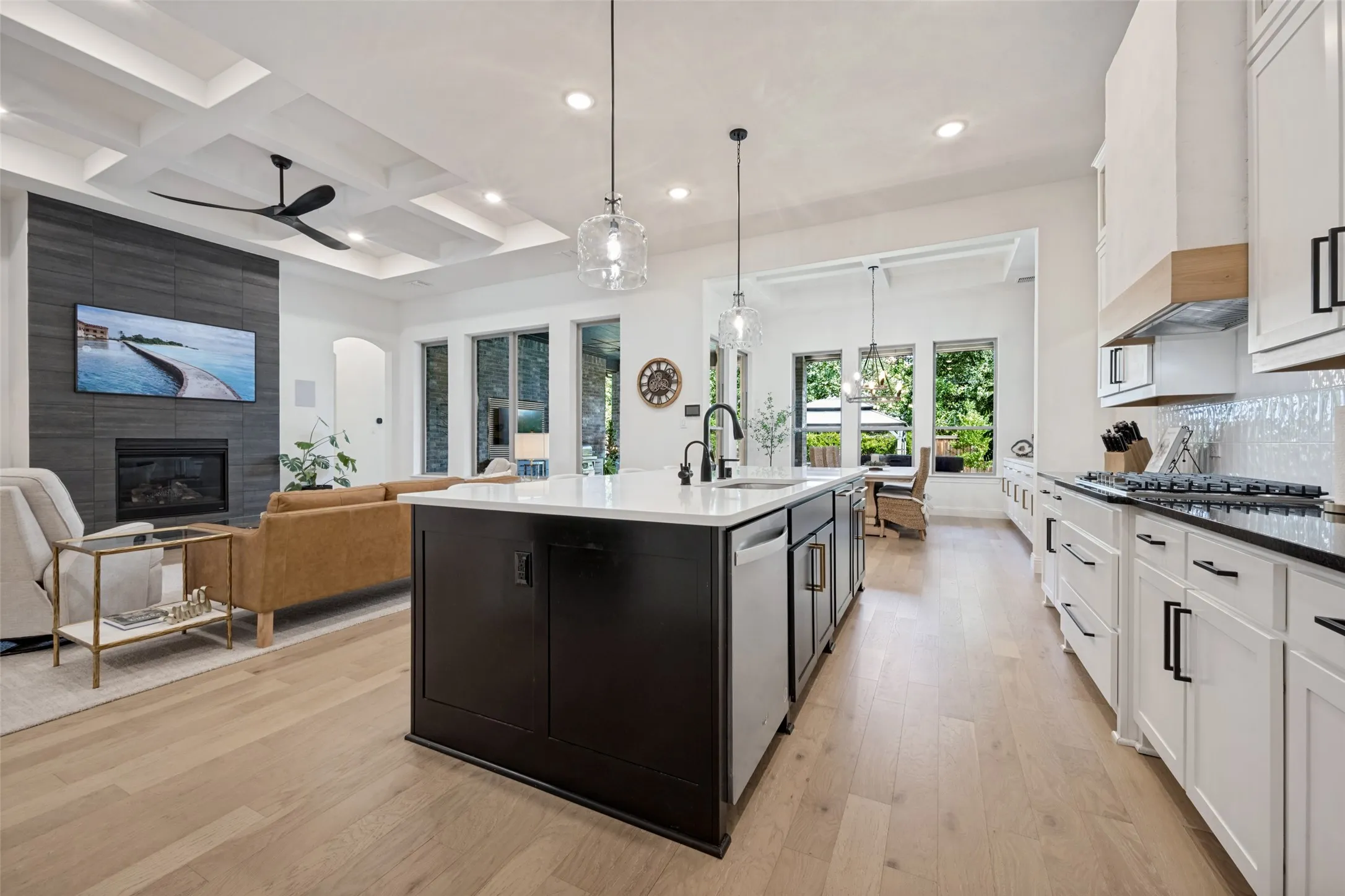 Light wood flooring throughout, coffered ceiling, built-in fireplace with dark tile surround, white kitchen cabinetry with dark hardware, and a center island with integrated appliances