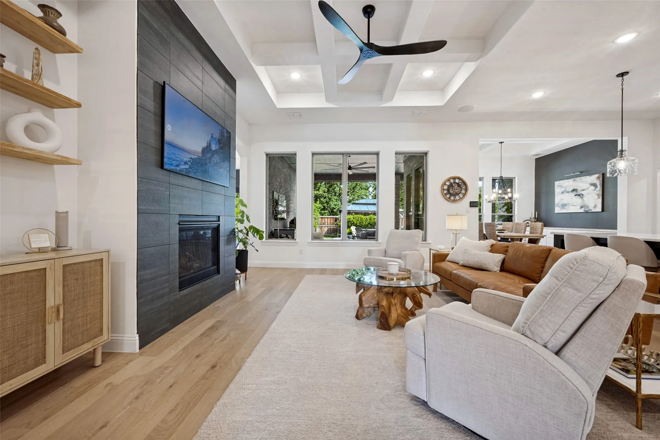 Living area featuring wood flooring, a contemporary fireplace with a dark tile surround, a coffered ceiling with recessed lighting, and multiple windows. Custom cabinetry and stunning floating shelves.