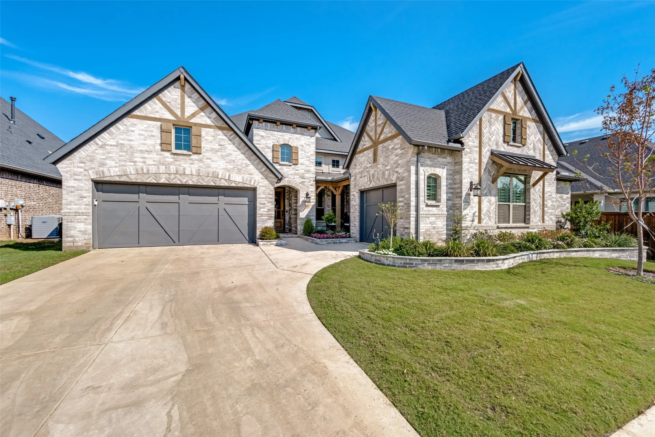 Stone exterior with dark trim accents, multiple gables, three attached garages, landscaped front yard, and a curved concrete driveway