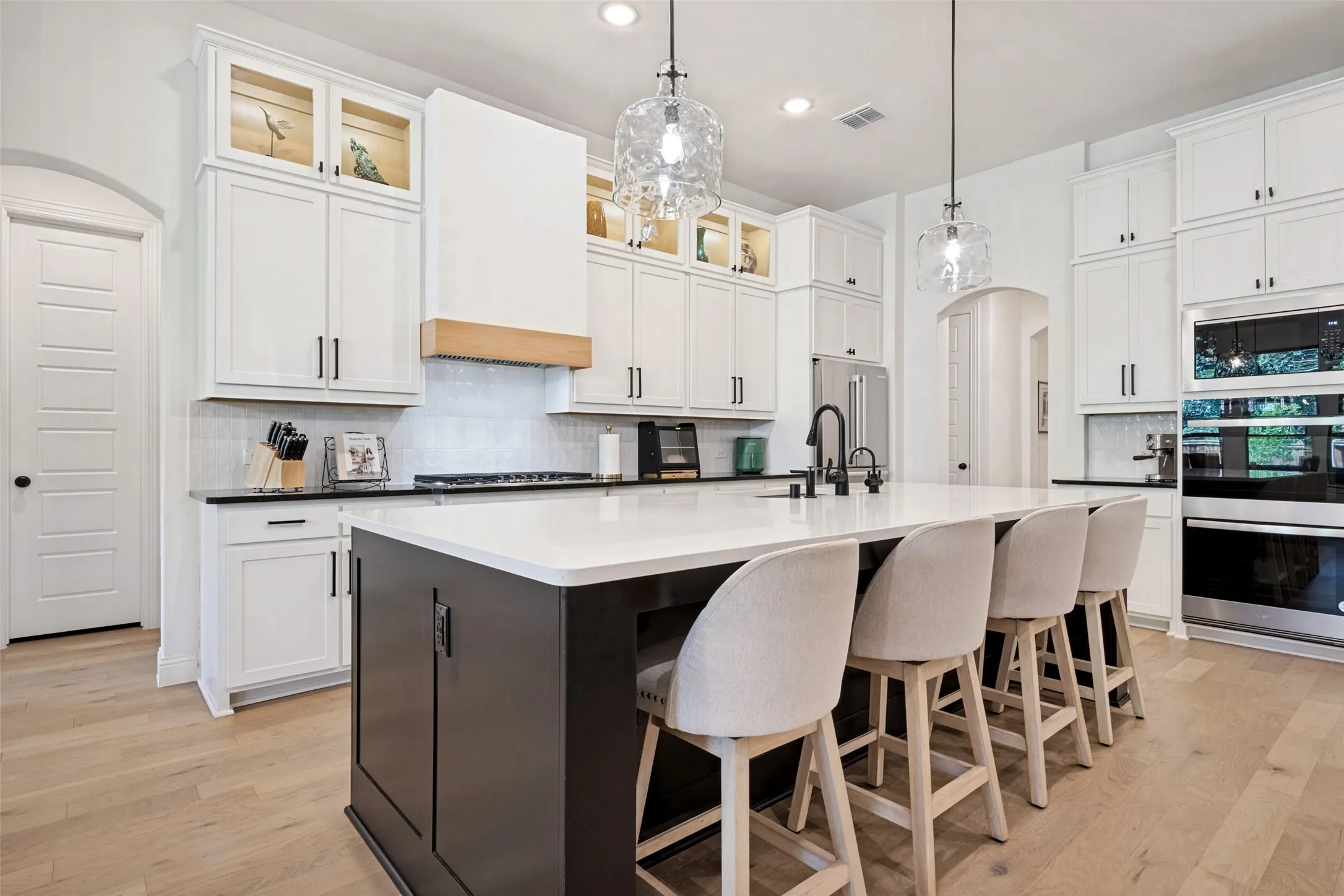 Gourmet kitchen featuring a large central island, white shaker-style cabinetry, light wood-finish flooring, and built-in stainless steel appliances. Custom range hood, lighted upper cabinets, filtered water and hot water spigot hard plumbed into kitchen island.