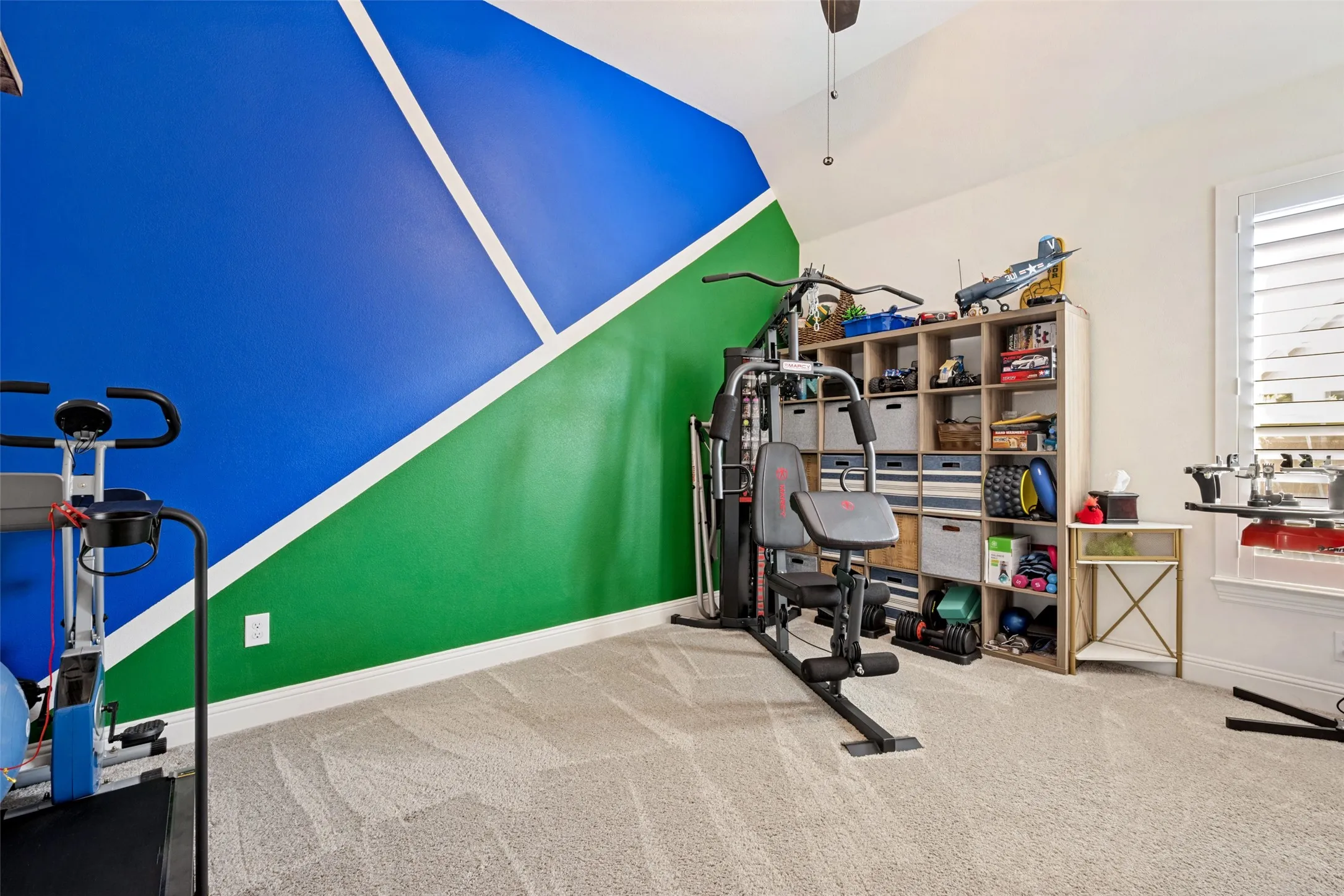 Vaulted ceiling, ceiling fan, window with plantation shutters, carpeted flooring, and decorative accent wall