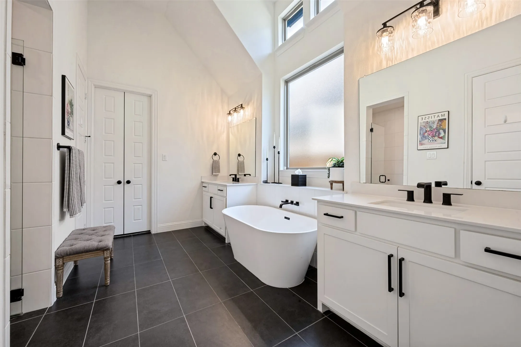Freestanding soaking tub, dual vanities with white countertops, dark tile flooring, and a large privacy window