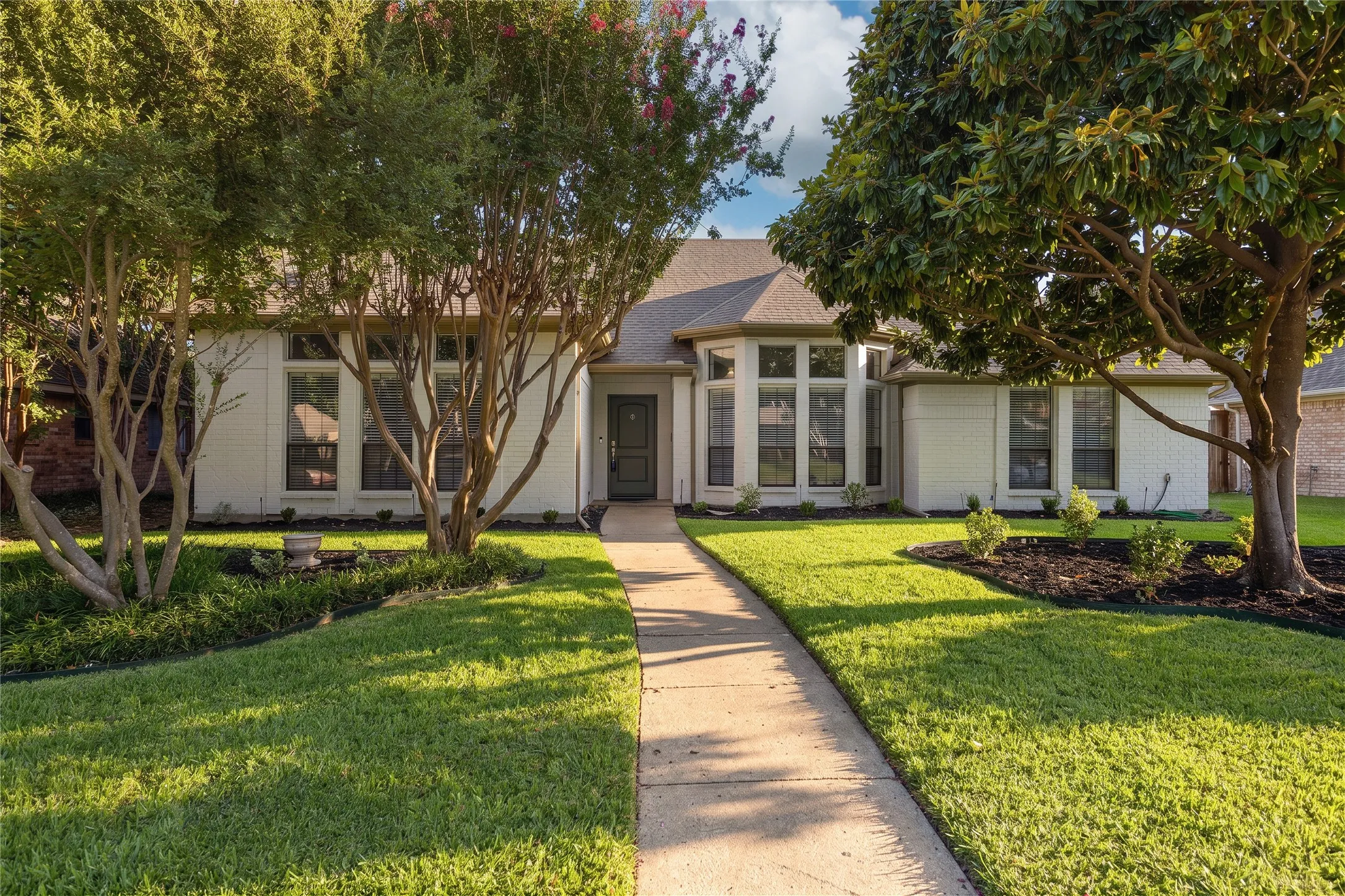 White exterior with a dark entry door, multiple large windows, a concrete walkway, and landscaped yard with mature trees