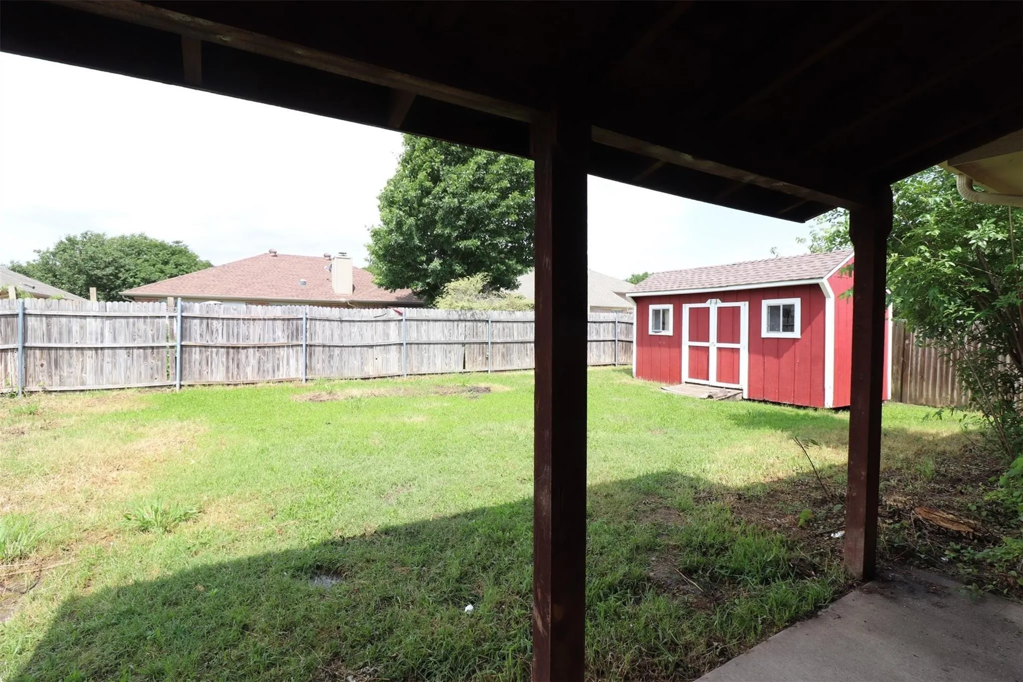 Expansive green space featuring a wooden privacy fence, a red storage shed with white trim, and a concrete patio area
