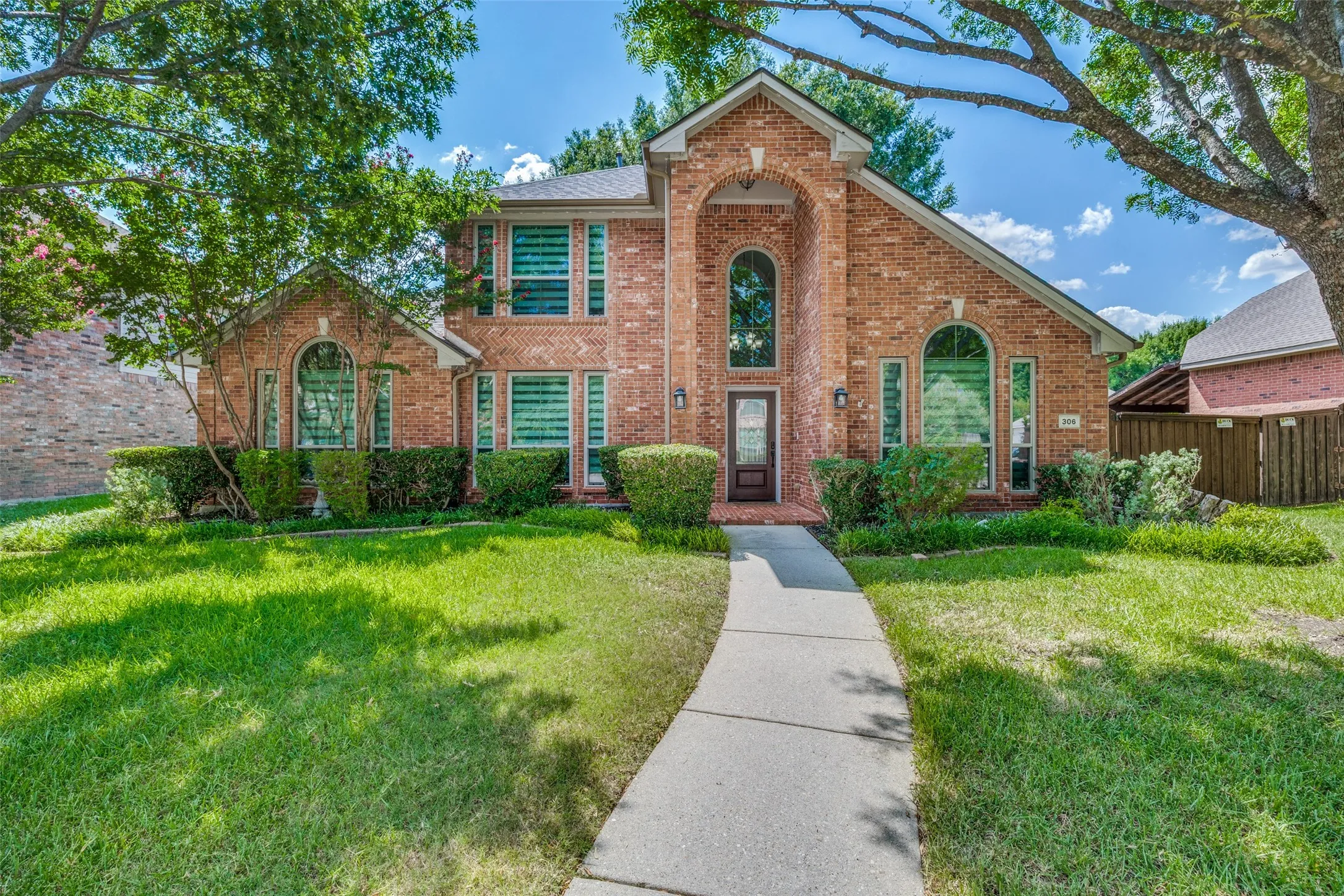 Brick exterior with arched windows, mature trees, a concrete pathway, and established foundation plantings