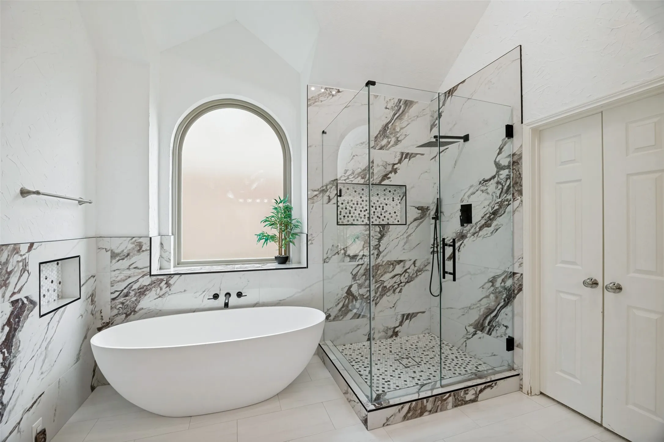 Freestanding soaking tub, glass-enclosed shower with rain head, marble-look tile accents, arched window, and dual panel doors