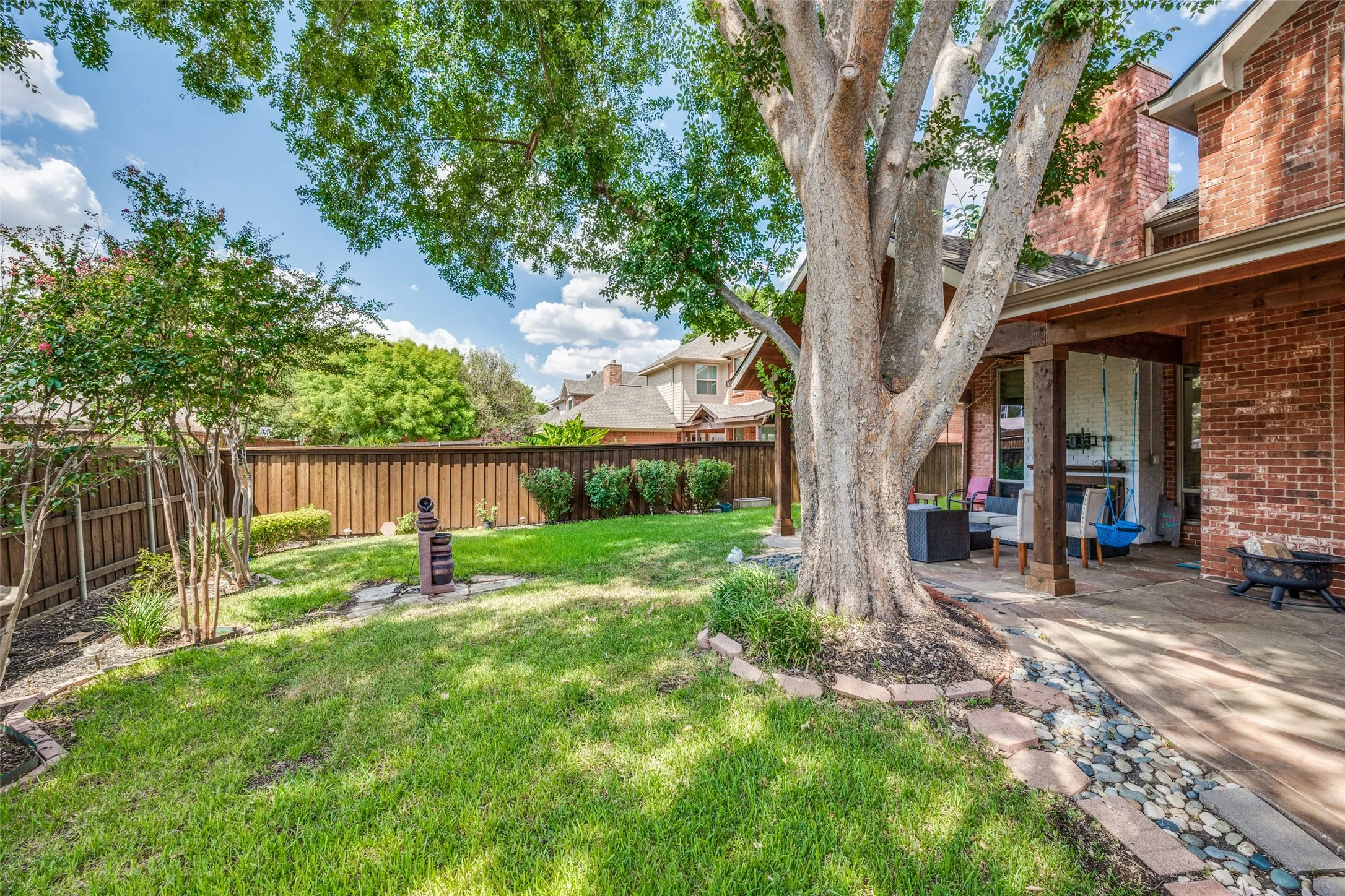 Expansive backyard featuring a manicured lawn, established perimeter fencing, mature trees, and a covered patio area with a stone-finish floor