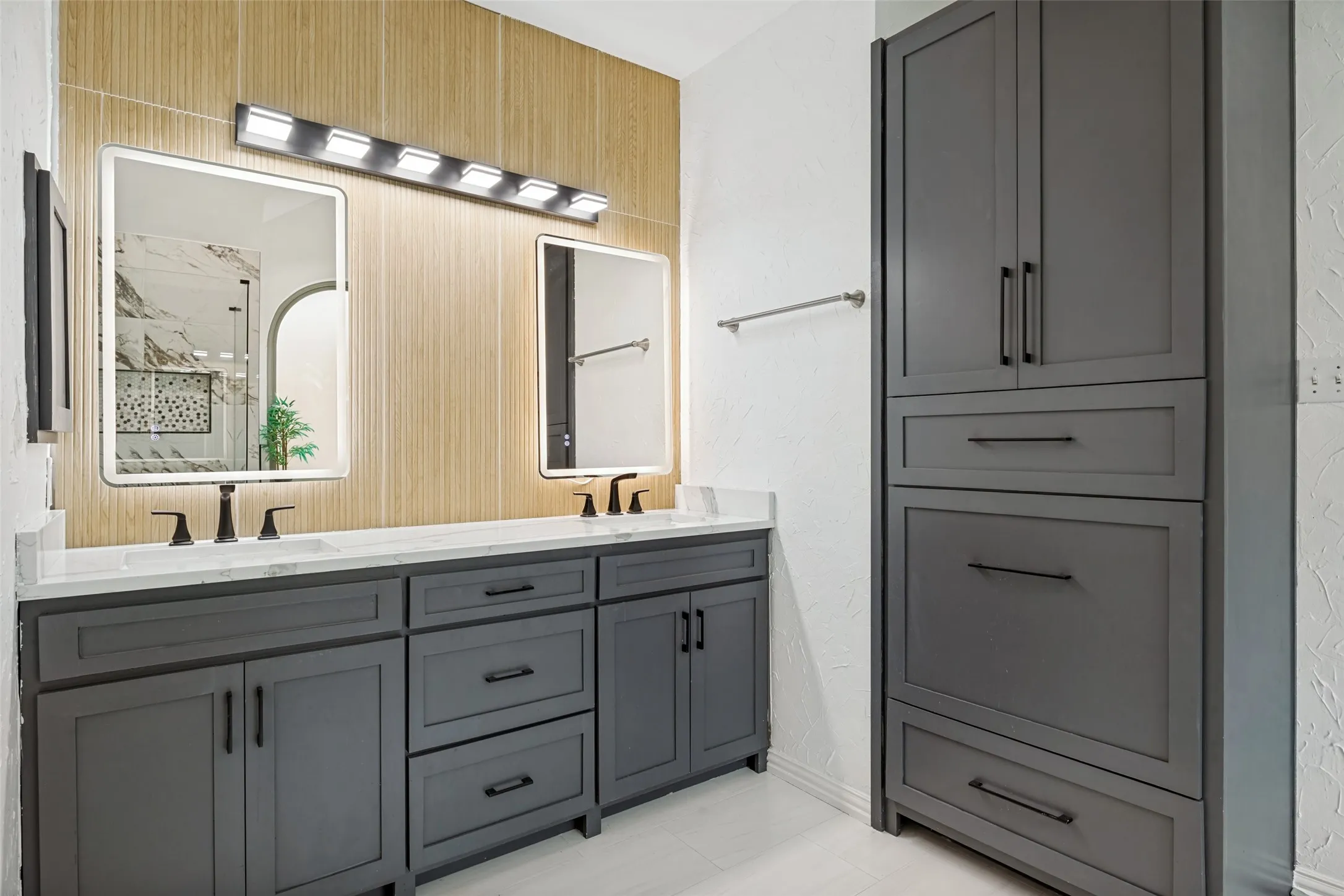 Dual rectangular mirrors with integrated lighting, wood-finish accent wall, double vanity with white countertops, dark gray cabinetry with matte black hardware, and an integrated tall storage cabinet