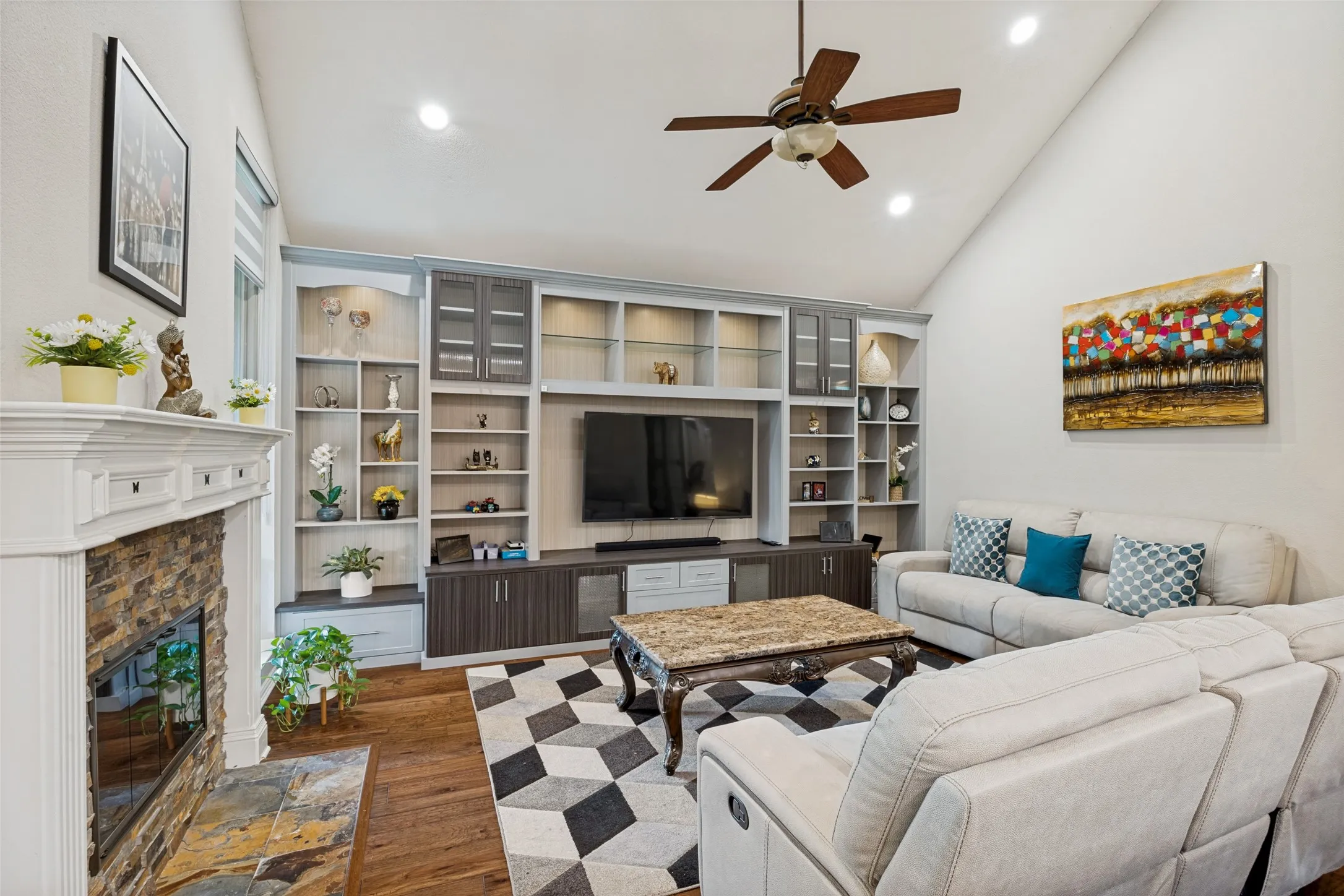 Vaulted ceiling, built-in entertainment center with shelving and cabinetry, fireplace with stone surround, wood-finish flooring, recessed lighting