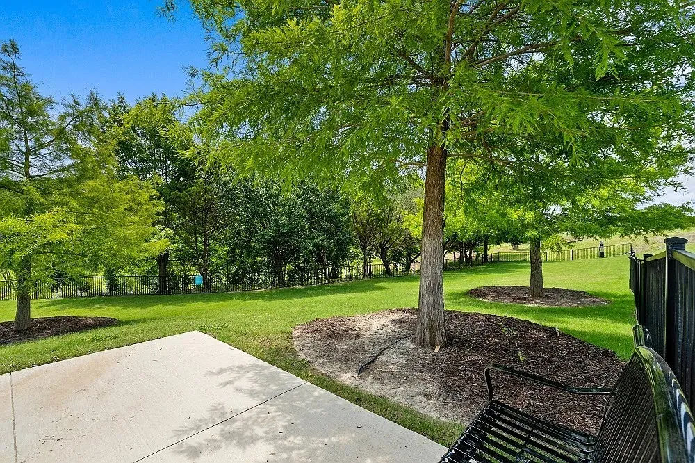 Concrete patio with a black metal fence, mature trees, and a grassy expanse