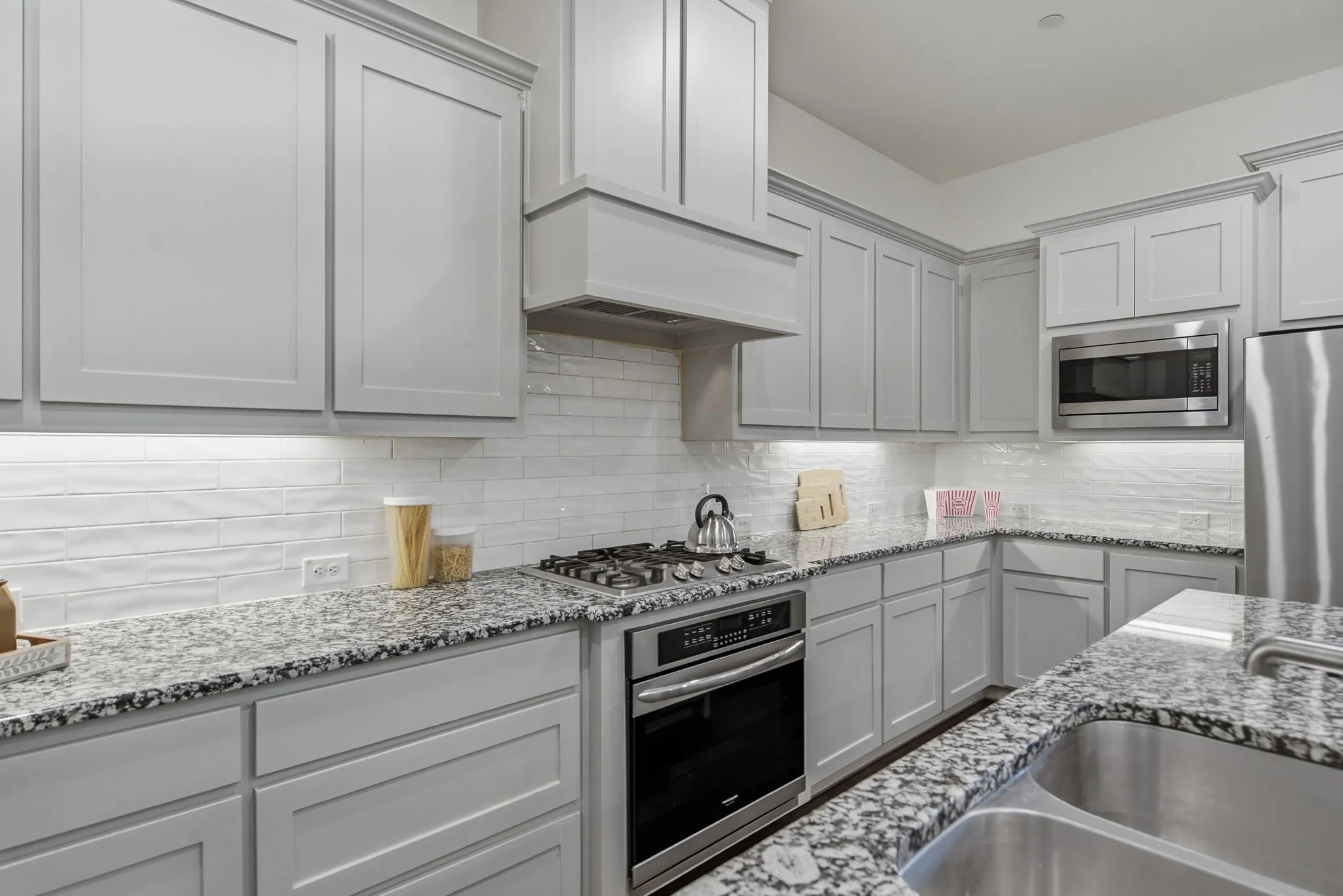 Modern kitchen featuring light gray cabinetry with crown molding, granite countertops, a stainless steel gas range with coordinating vent hood, and a built-in microwave