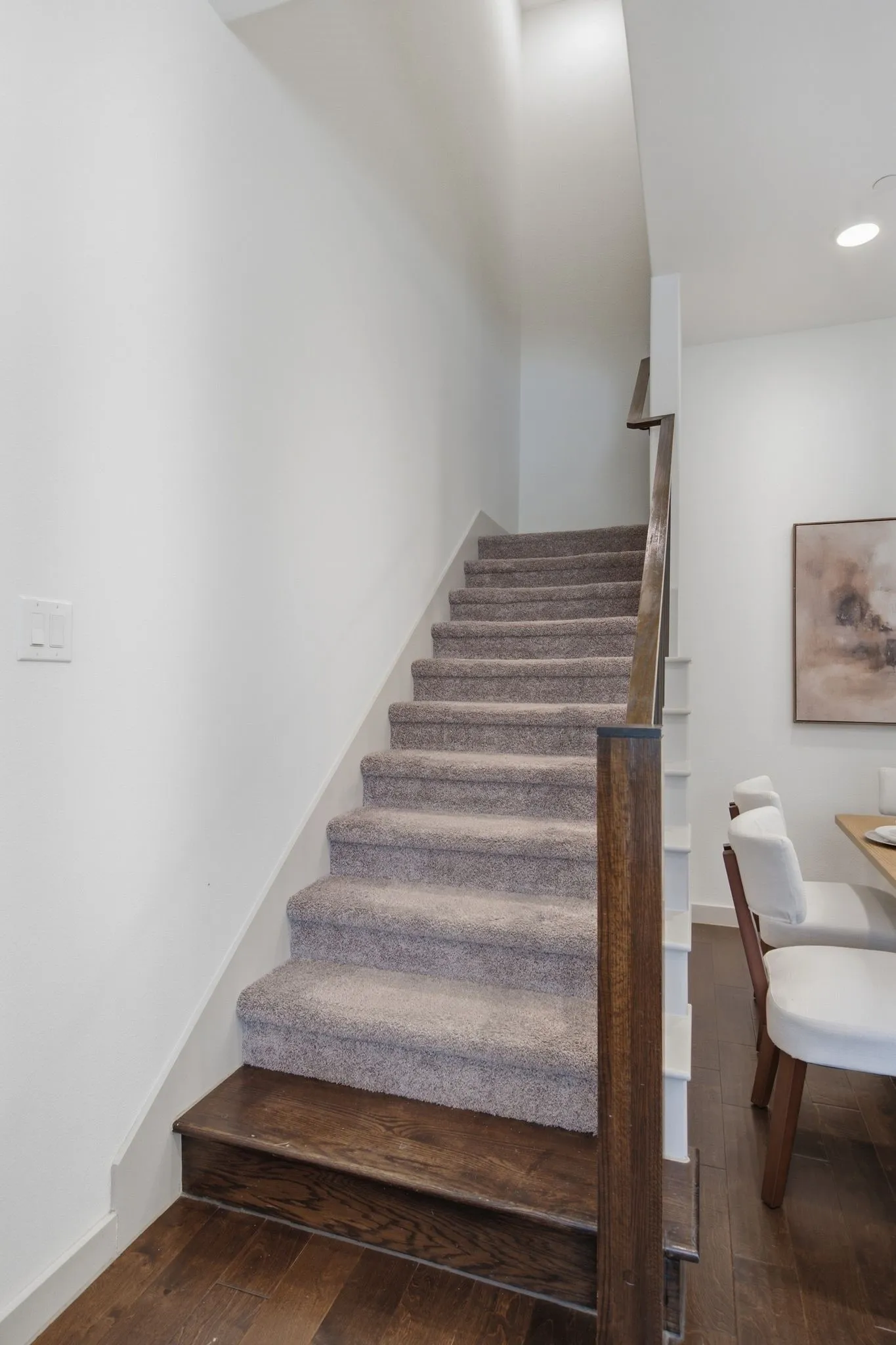 Wood-finish flooring leading to a carpeted staircase with dark wood-finish newel posts and handrail, set against white walls