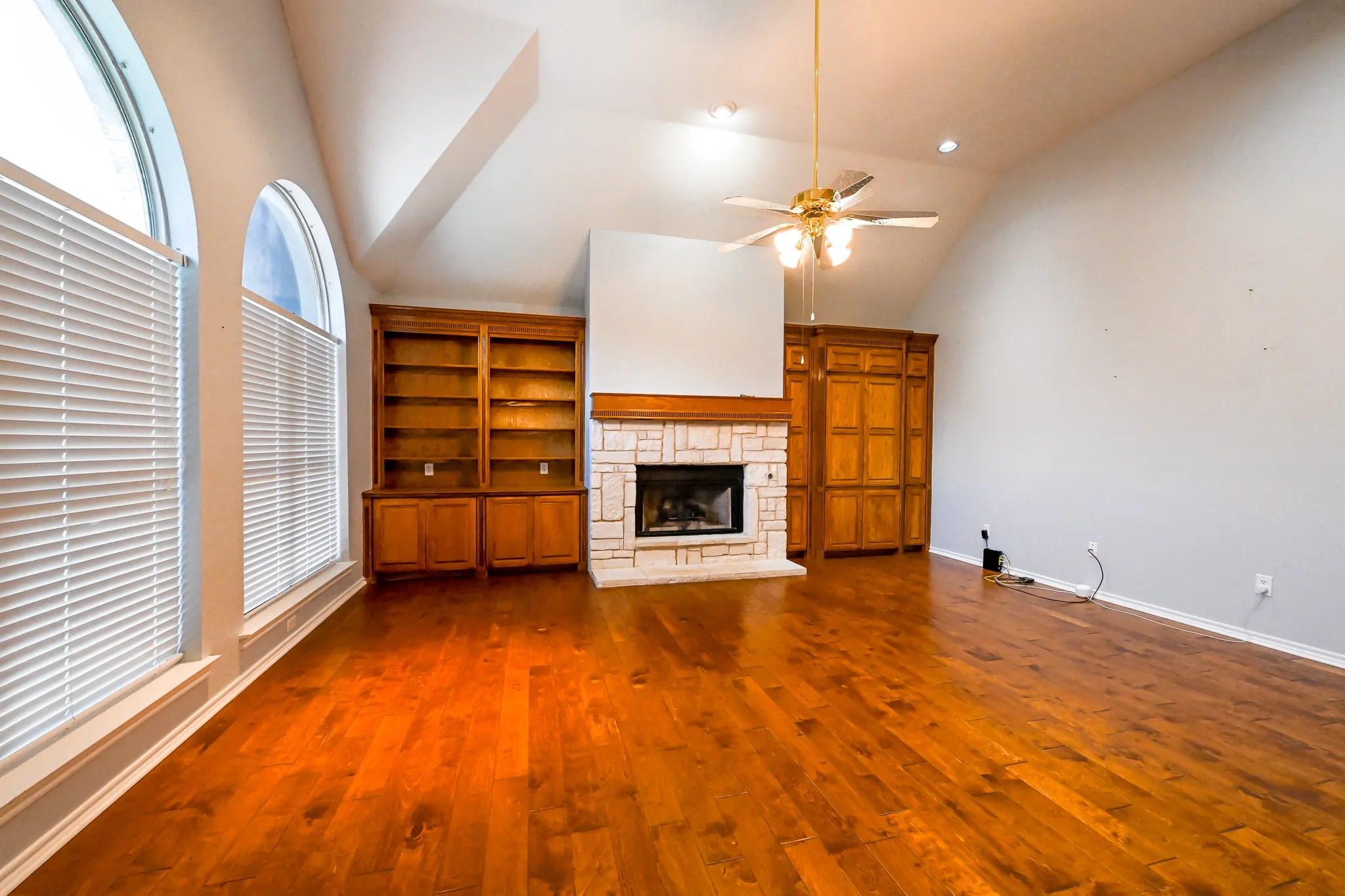 Vaulted ceiling, wood-finish flooring, stone-clad fireplace, built-in shelving, and arched windows