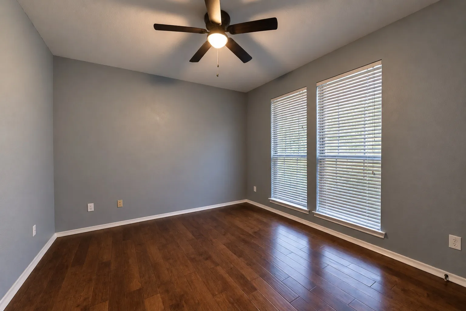 Wood-finish flooring, two windows with blinds, ceiling fan with integrated lighting, and gray painted walls