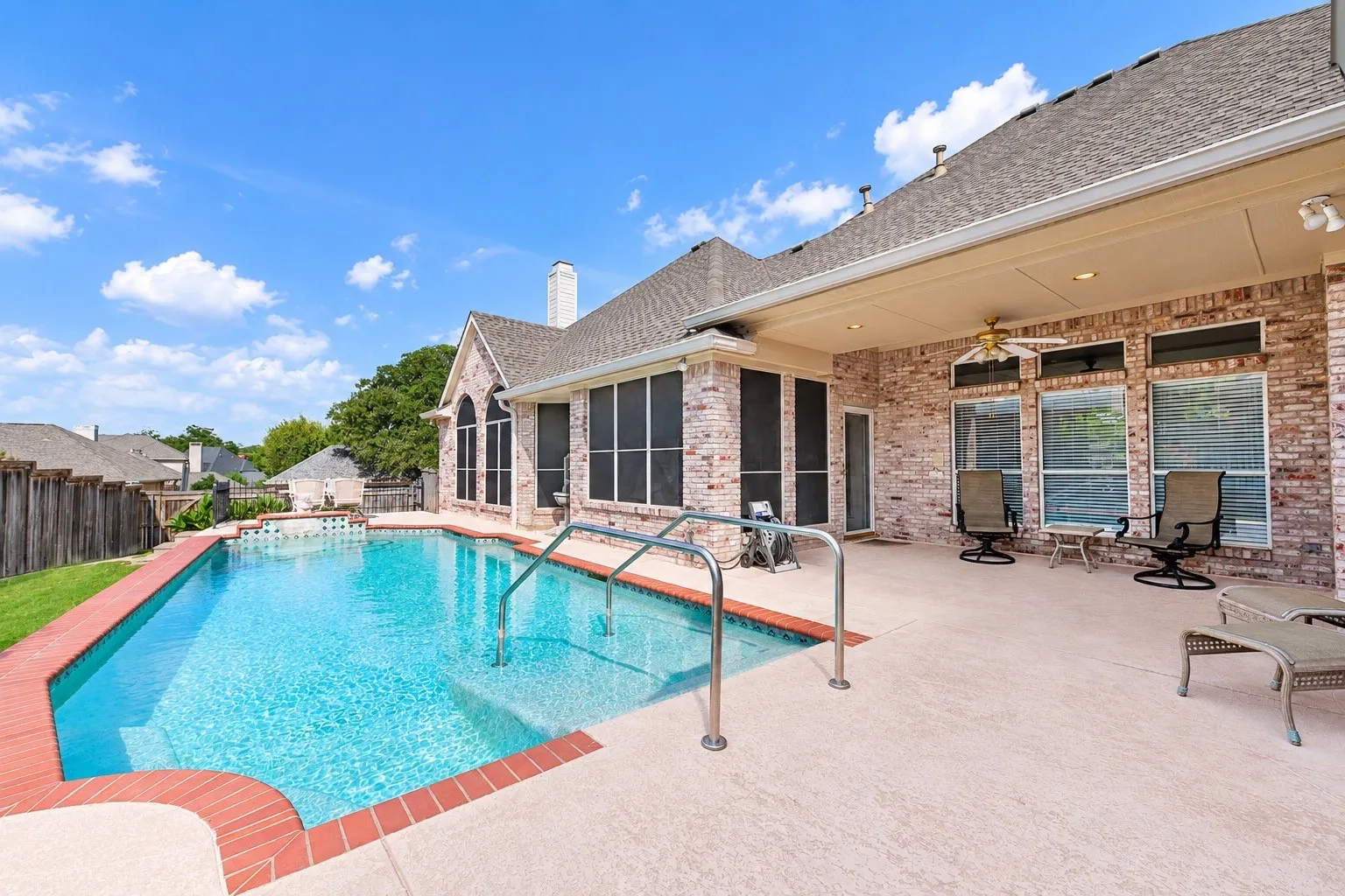 Private swimming pool with integrated steps, expansive concrete patio, covered outdoor living space with ceiling fan, and brick exterior
