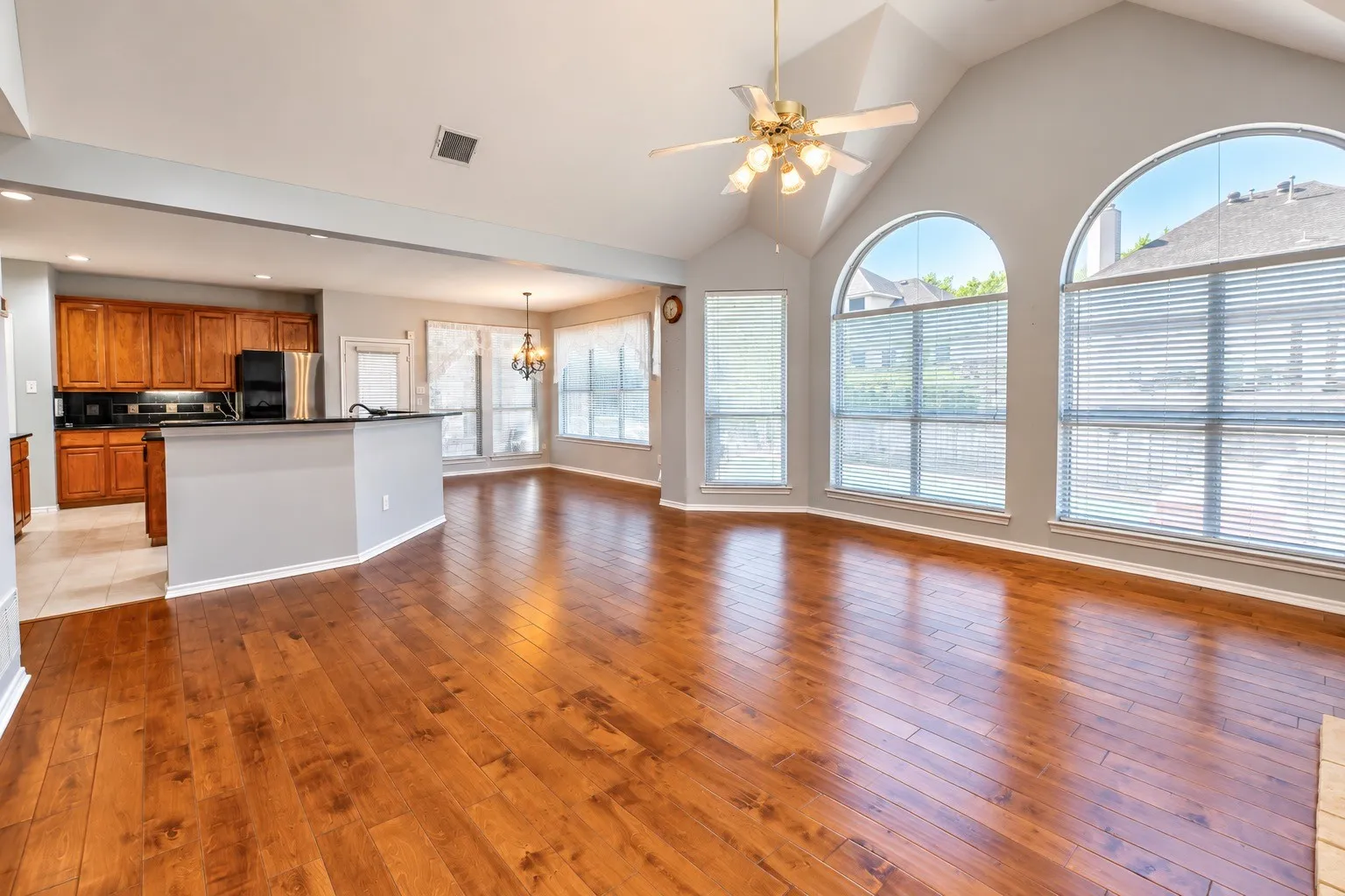 Vaulted ceiling with ceiling fan, arched windows with blinds, wood-finish flooring, kitchen with wood cabinetry and black appliances, and a breakfast bar