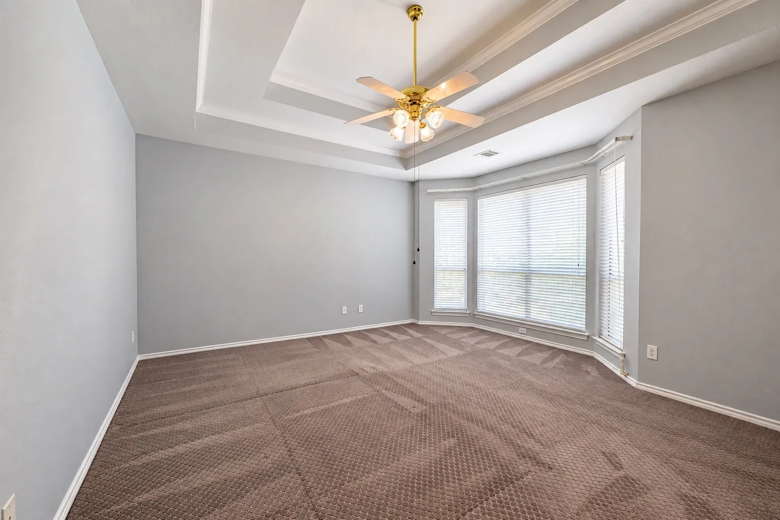 Tray ceiling with crown molding, ceiling fan with integrated lighting, bay windows with blinds, and neutral carpet flooring