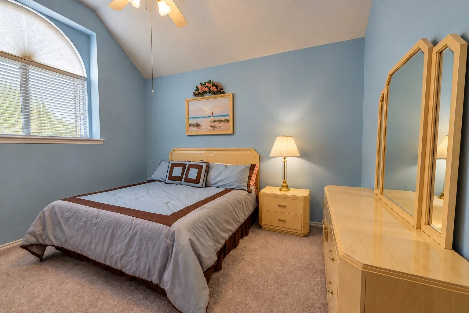 Vaulted ceiling with a ceiling fan, arched window with horizontal blinds, neutral carpet, and a light wood-finish dresser with a three-panel mirror