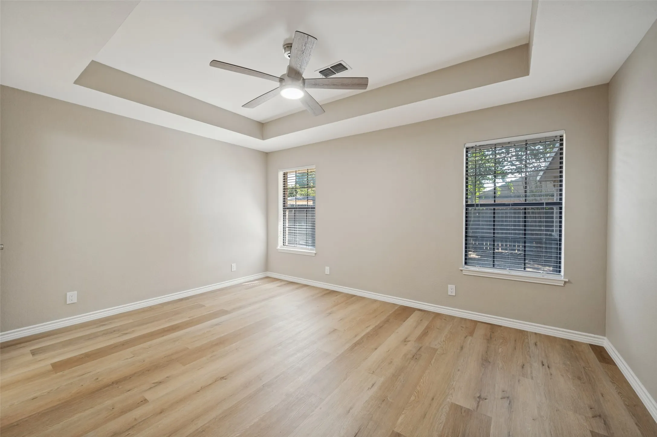 Tray ceiling with a central ceiling fan, LVP flooring, and two windows with blinds