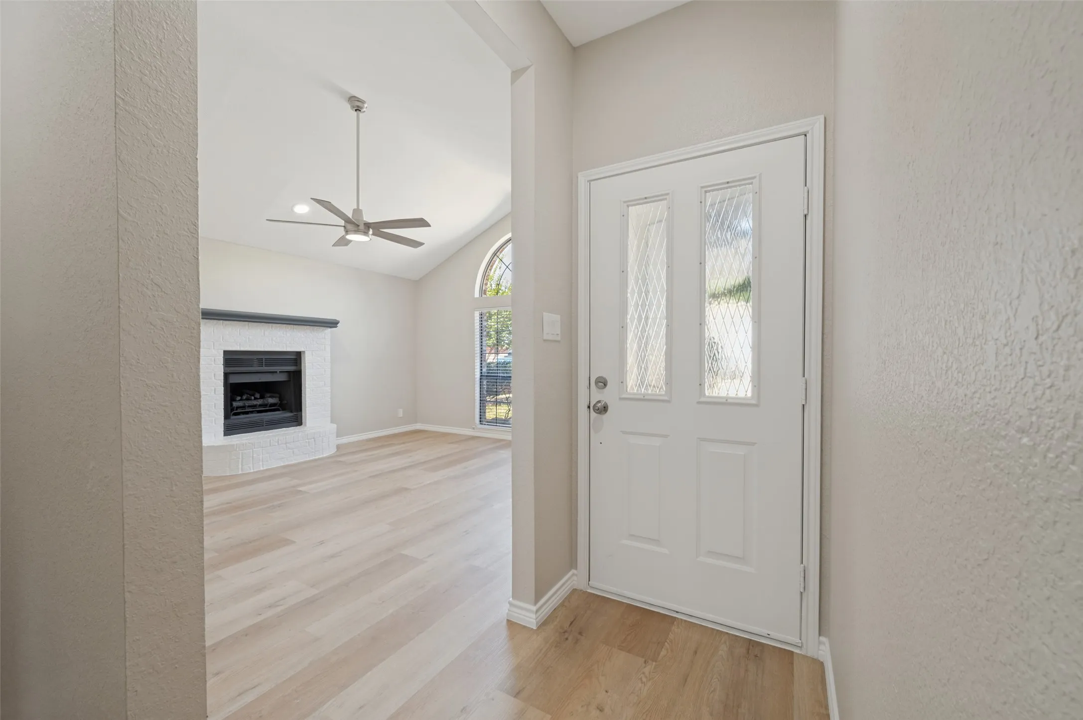 Light-colored LVP flooring, vaulted ceiling, white tiled fireplace with dark mantel, arched window, and a white entry door with decorative glass panels