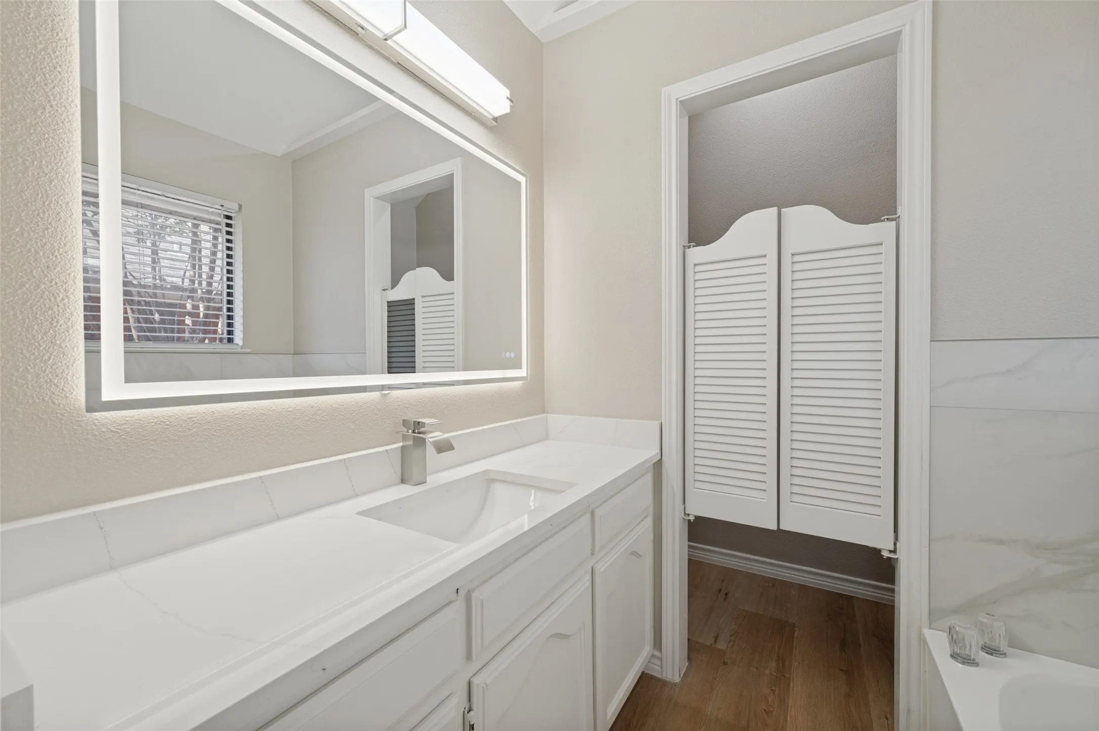 Bathroom vanity featuring an integrated sink, brushed nickel faucet, large illuminated mirror, and white  cabinetry