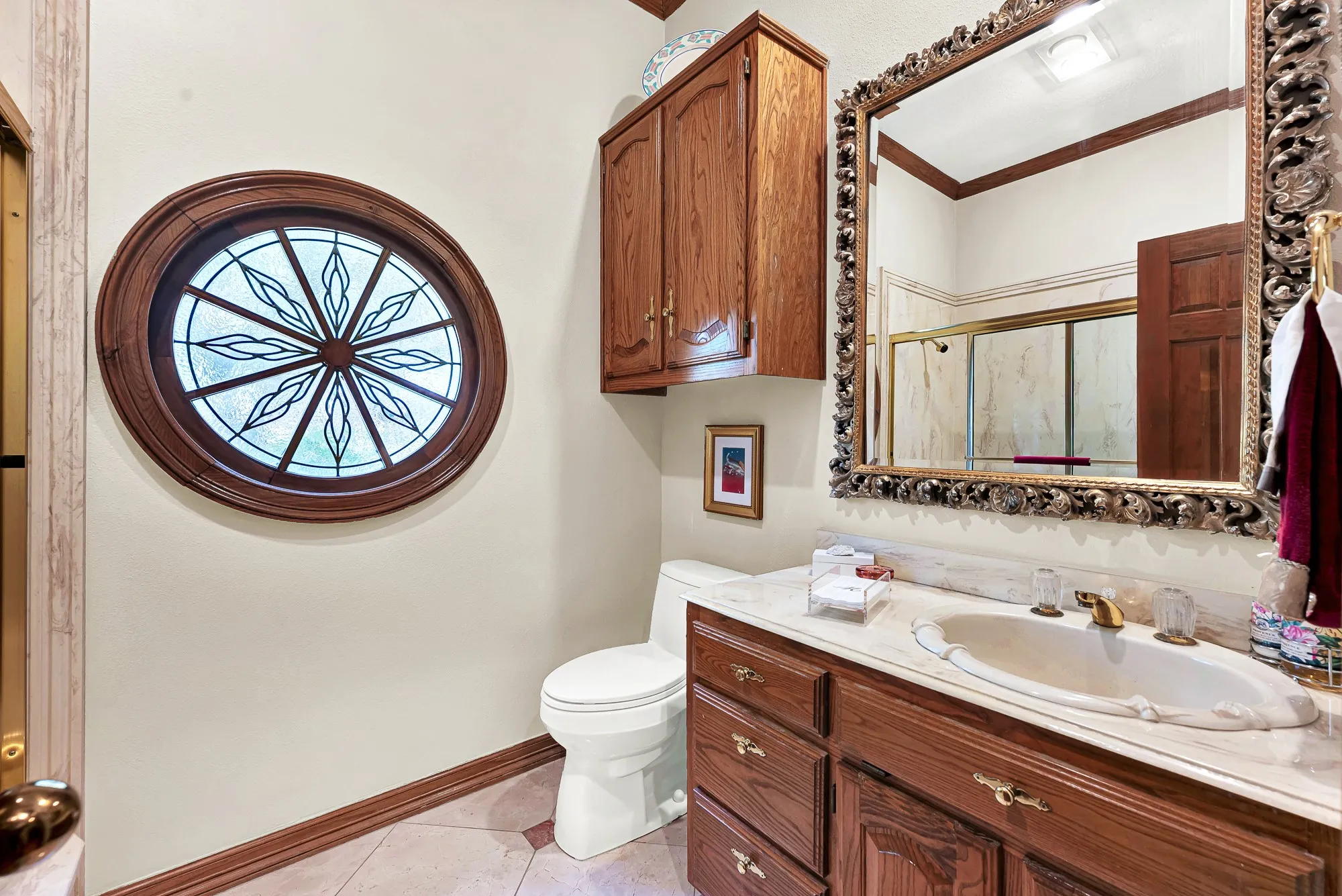 Bathroom on first floor showcasing custom wood cabinetry, a beautiful vanity, ornate carved mirror, and distinctive round leaded-glass window framed in rich wood.