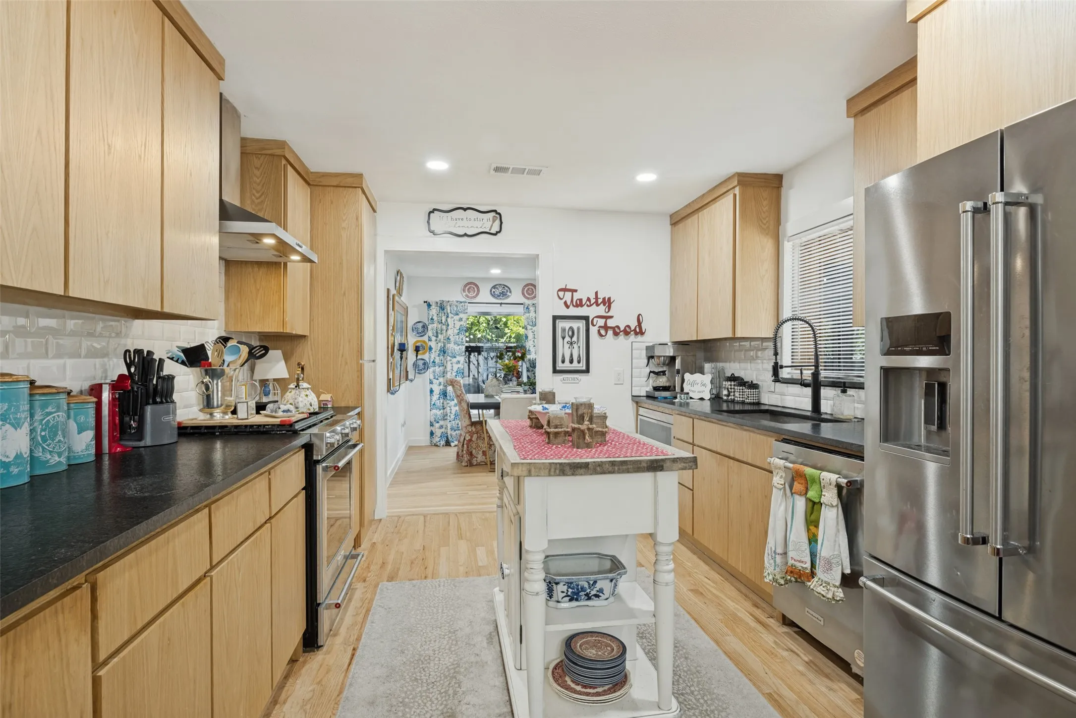 Kitchen featuring light wood-finish cabinetry, dark countertops, a subway tile backsplash, stainless steel appliances, and wood-finish flooring