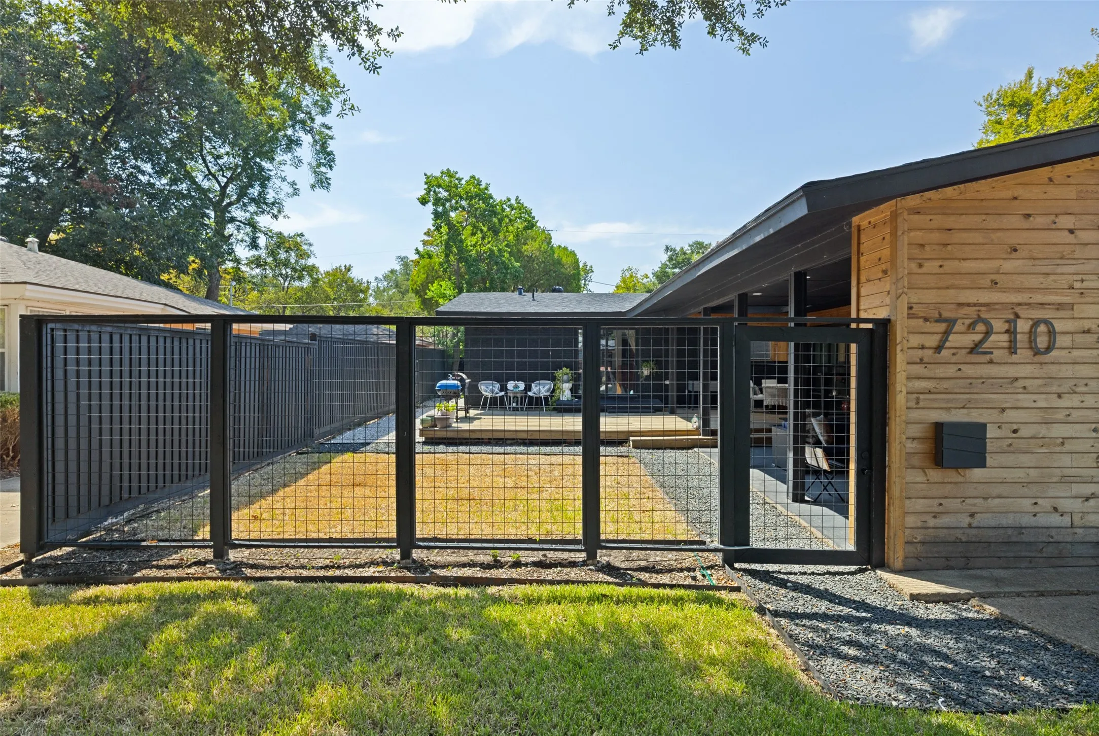 Horizontal wood-plank siding with exterior numbering, modern black mailbox, black metal fencing, and a gravel pathway