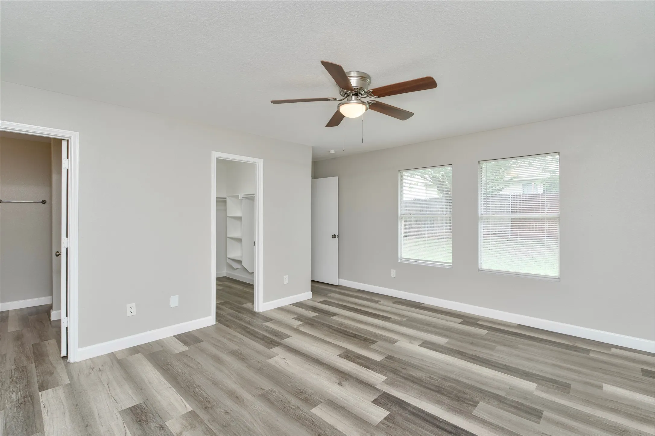 Spacious room featuring wood-finish flooring, light gray walls, two windows, and a ceiling fan with integrated lighting