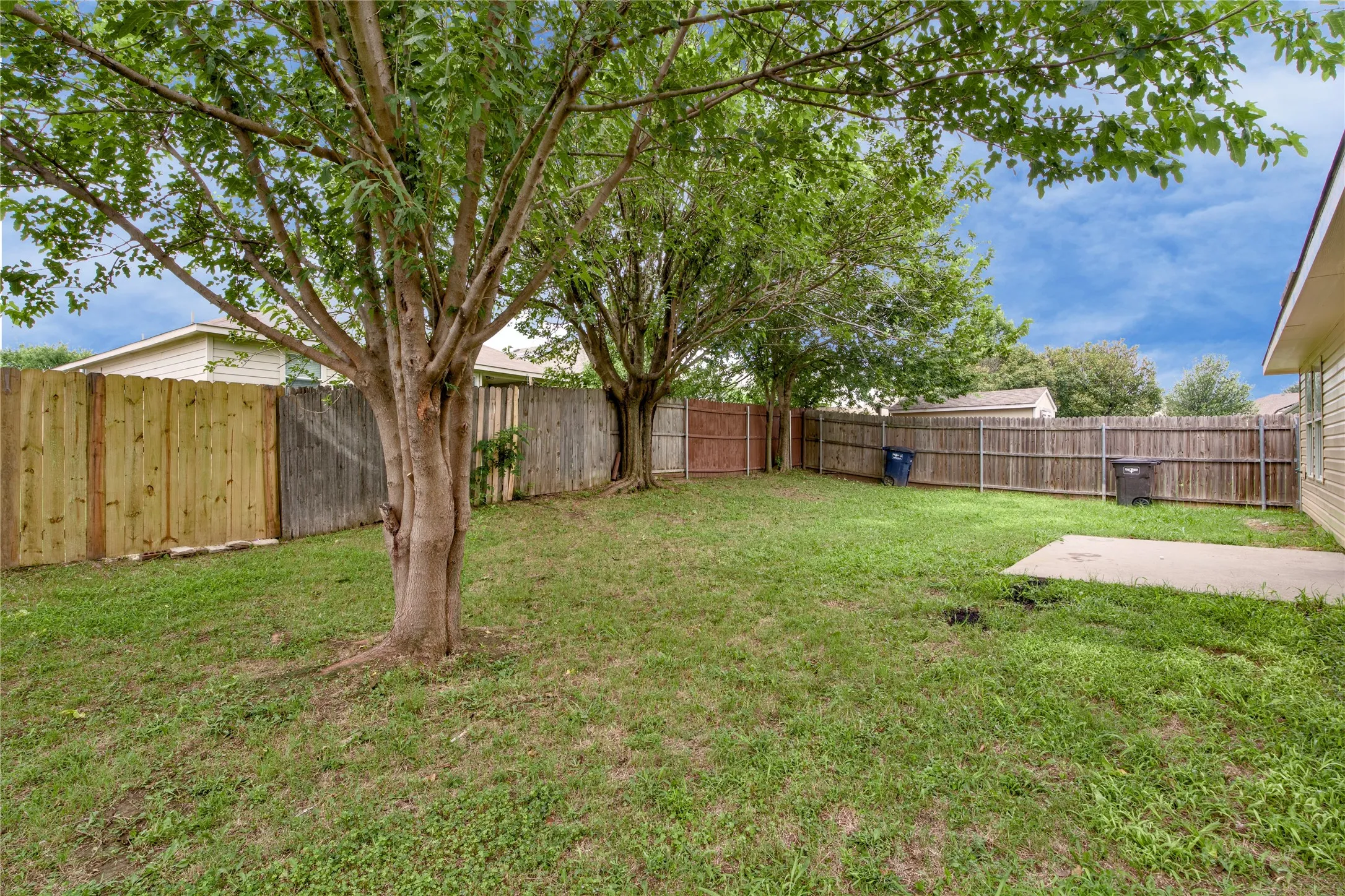 Expansive grassy yard with mature trees, wood fencing, and a concrete patio