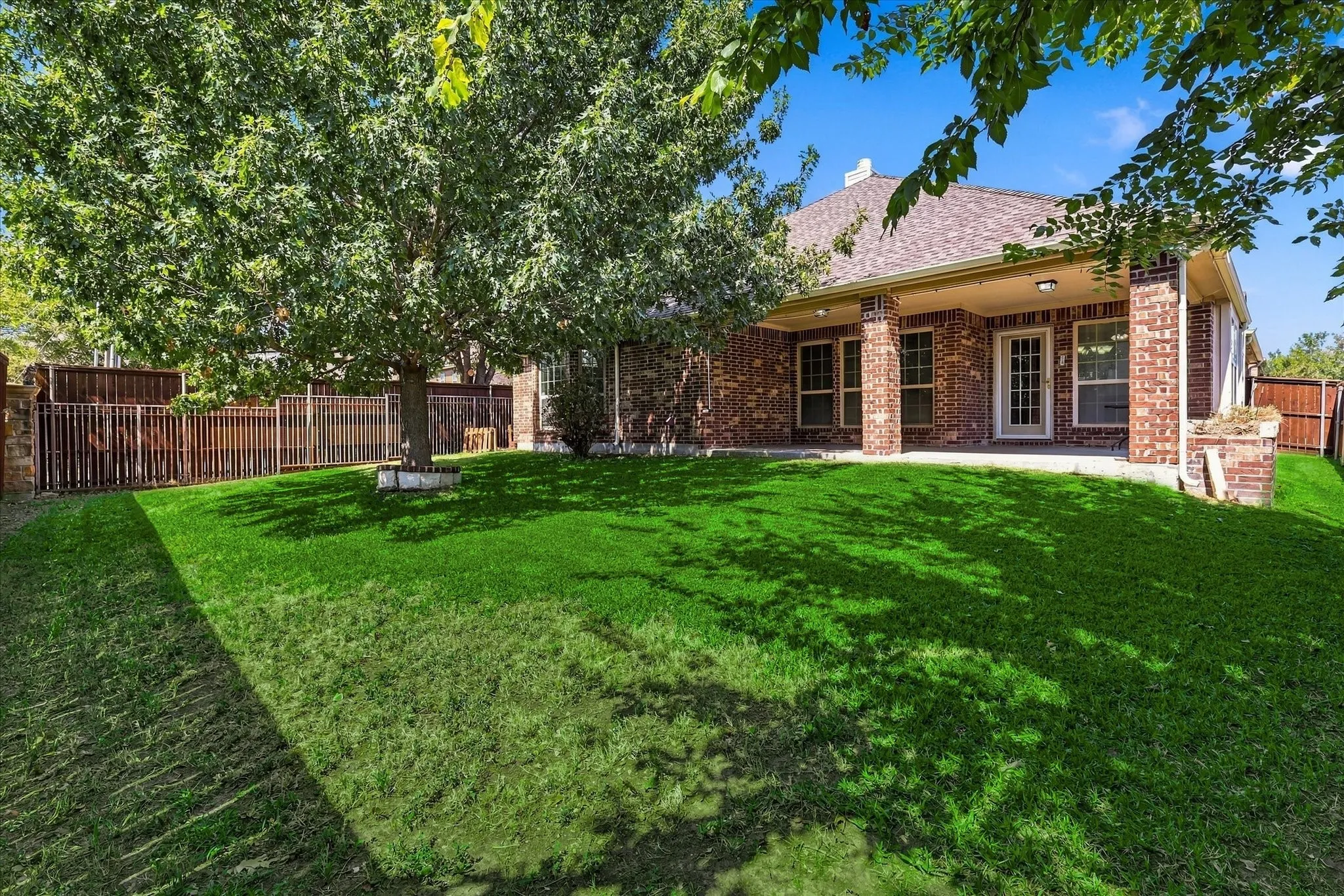 Expansive backyard featuring a covered patio with brick columns, a mature shade tree, a privacy fence, and a glass-paneled exterior door