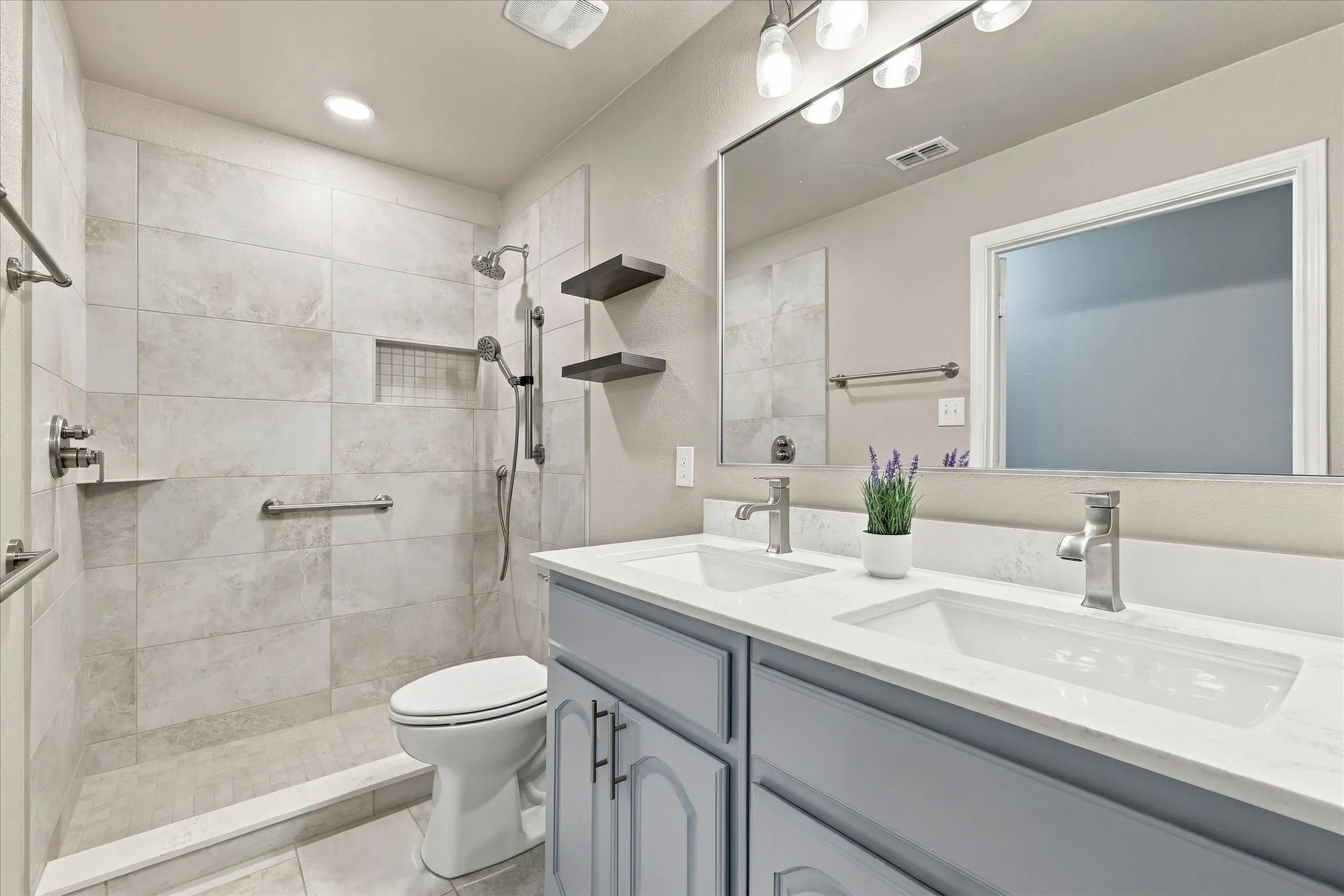Walk-in shower with tiled walls and floor, dual vanity with undermount sinks, and floating shelves