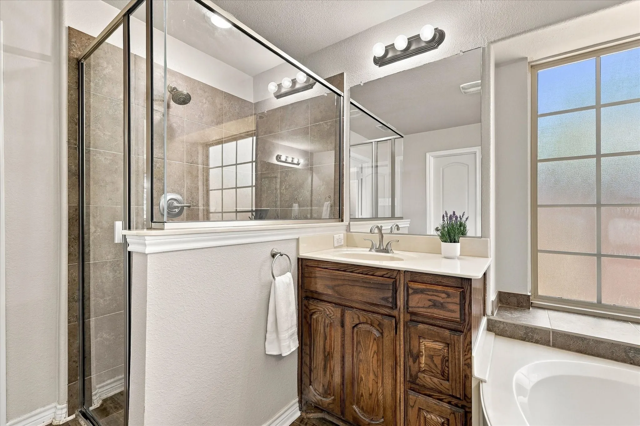 Glass-enclosed shower with tile surround, wood-finish vanity with countertop, large wall mirror, and window with frosted panes