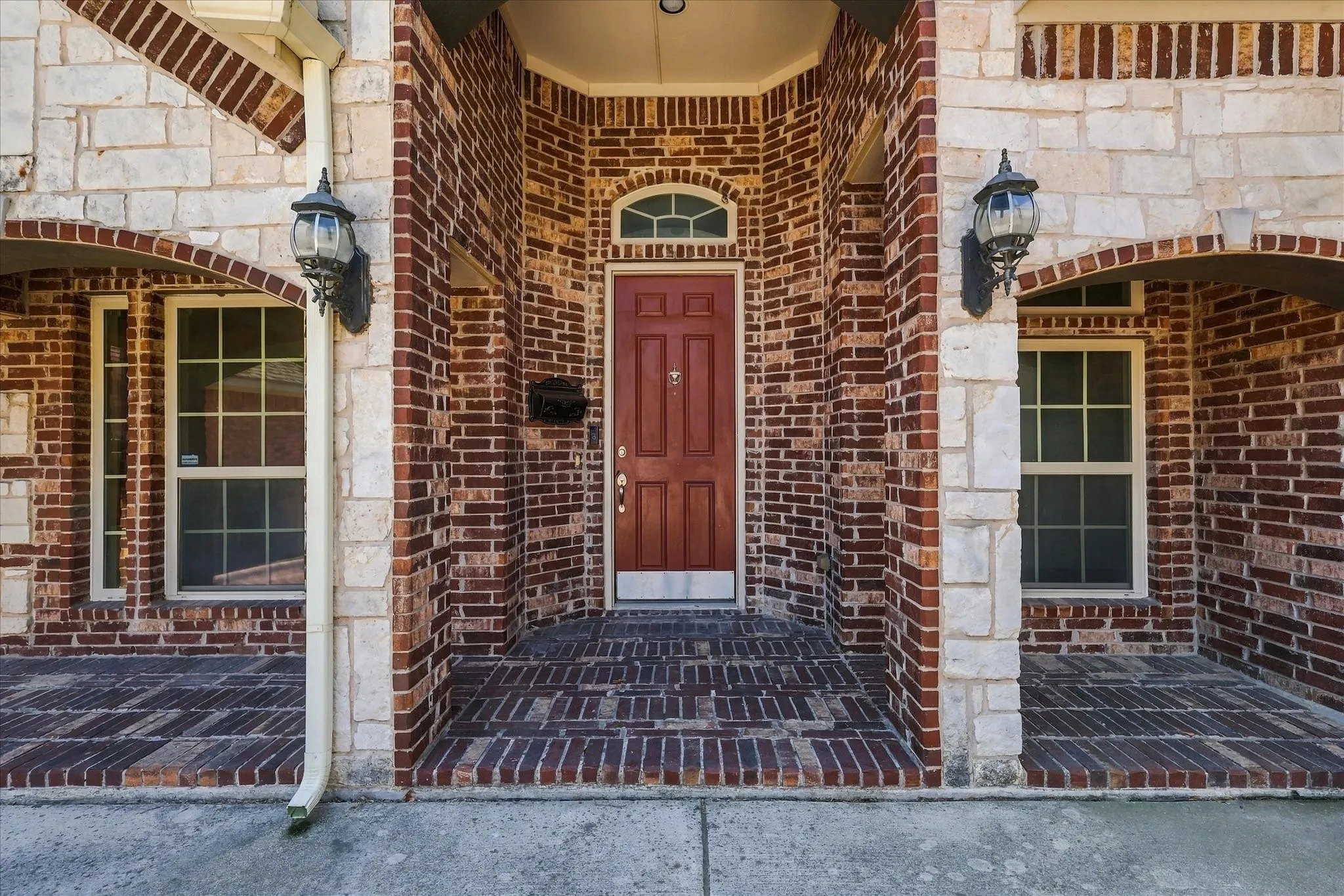 Brick and stone facade featuring a red entry door with an arched transom, twin wall-mounted lanterns, and a brick-patterned porch