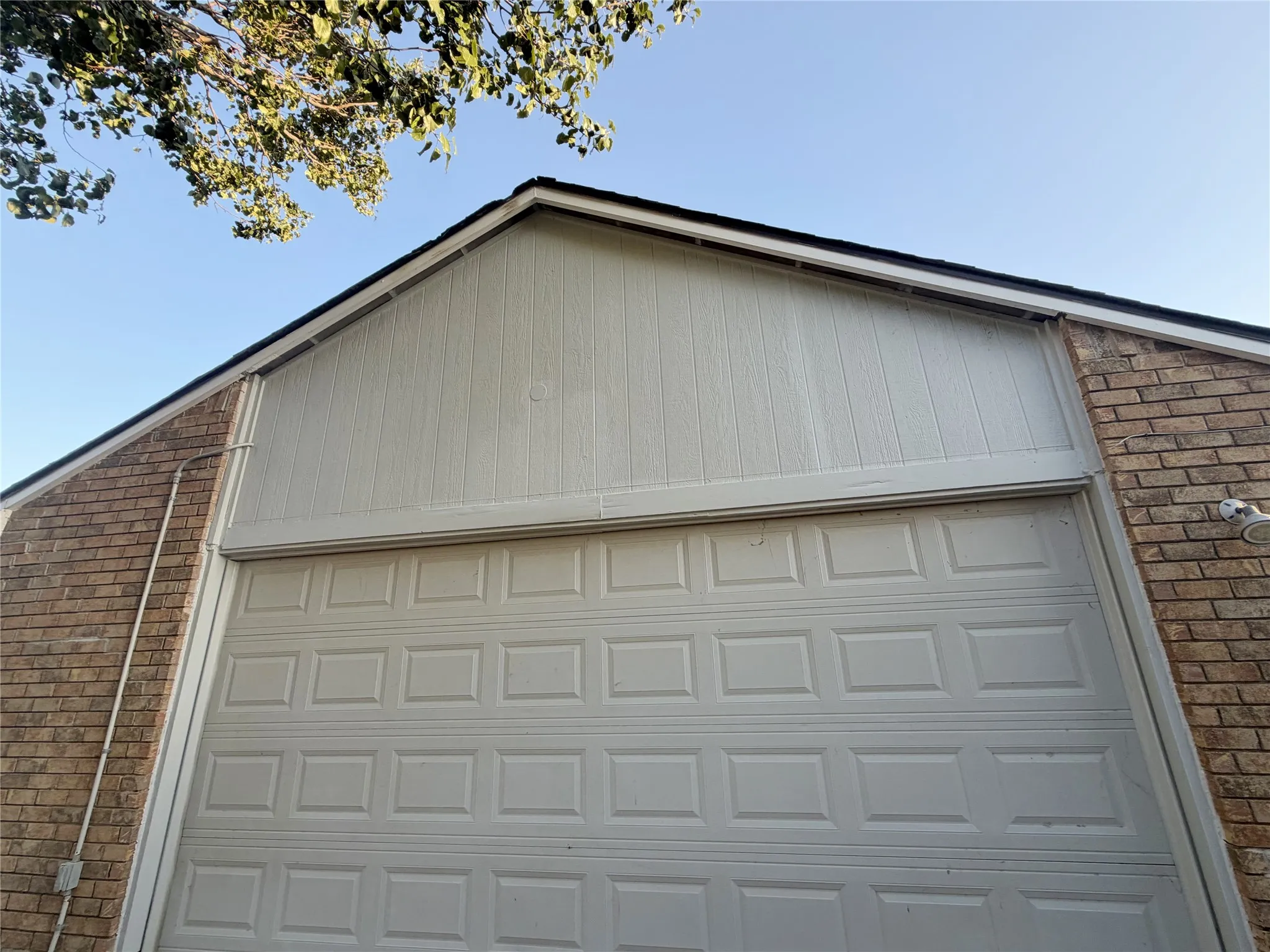 Attached garage with panel door, brick facade, and vertical siding on the gable
