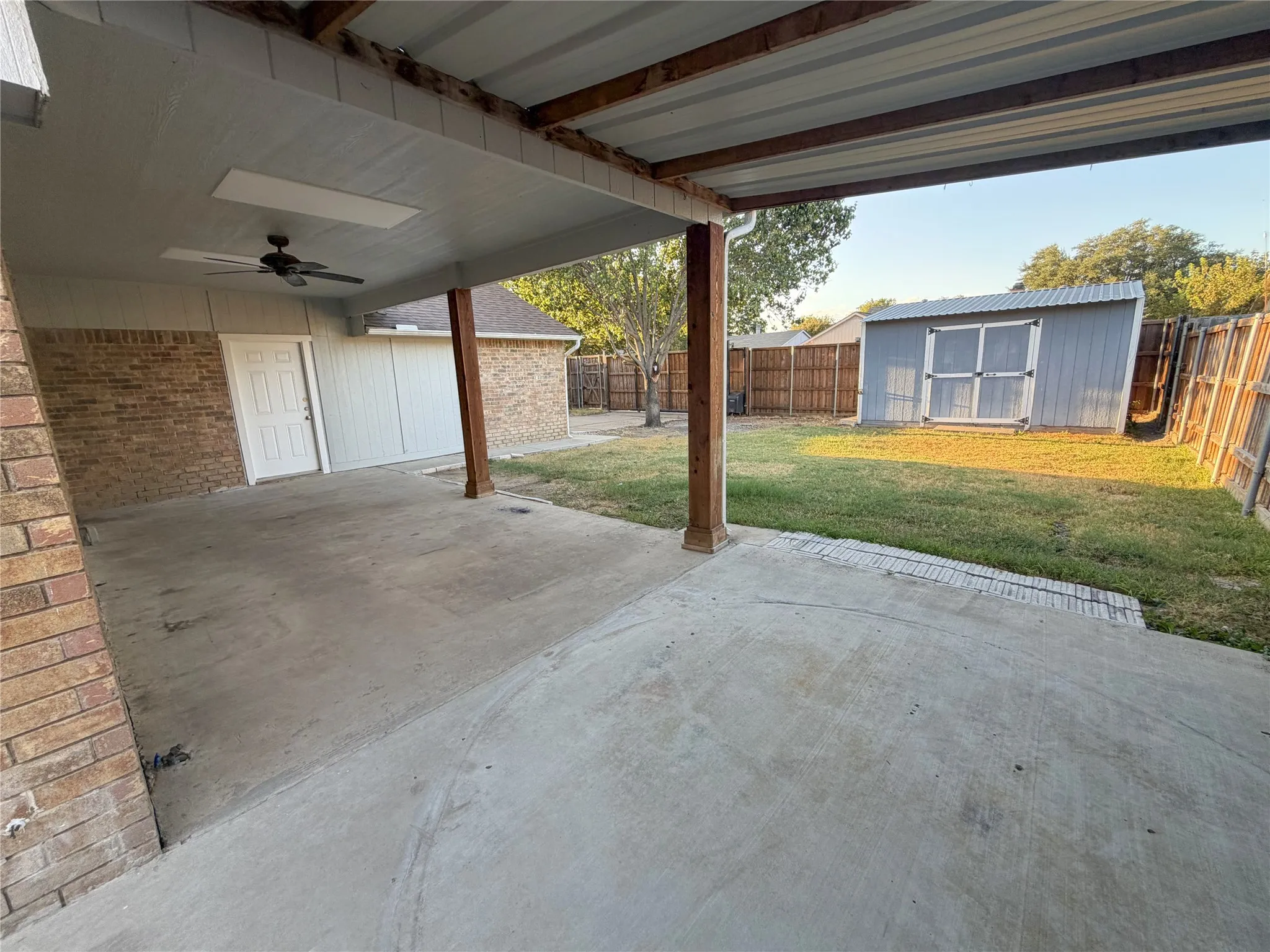 Covered concrete patio with ceiling fan, brick exterior, wood fence, and storage shed
