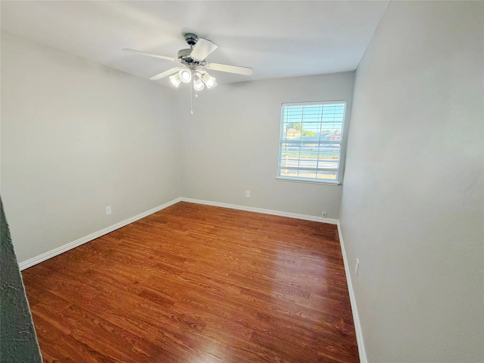 Wood-finish flooring, white baseboards, a window with blinds, and a ceiling fan with integrated lighting