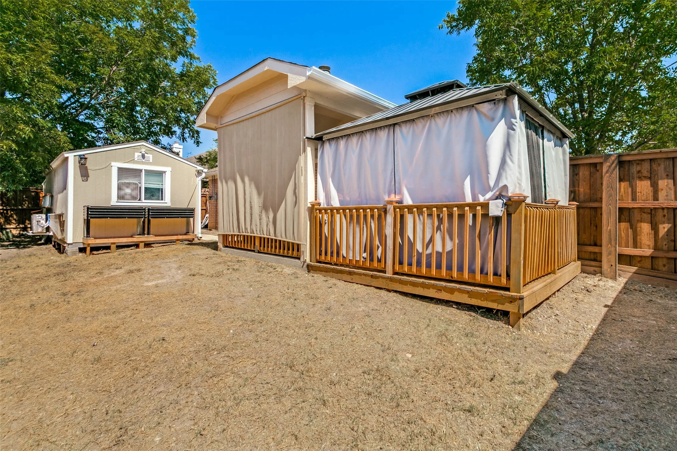 Gazebo with privacy drapes and a wood-railed deck, accompanied by a detached structure with a window and siding