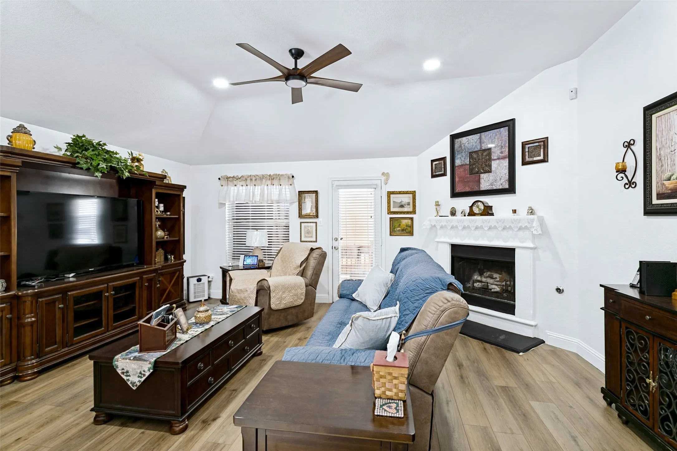 Vaulted ceiling with recessed lighting and a ceiling fan, fireplace with a decorative mantel, wood-finish flooring, and a glass-paneled door