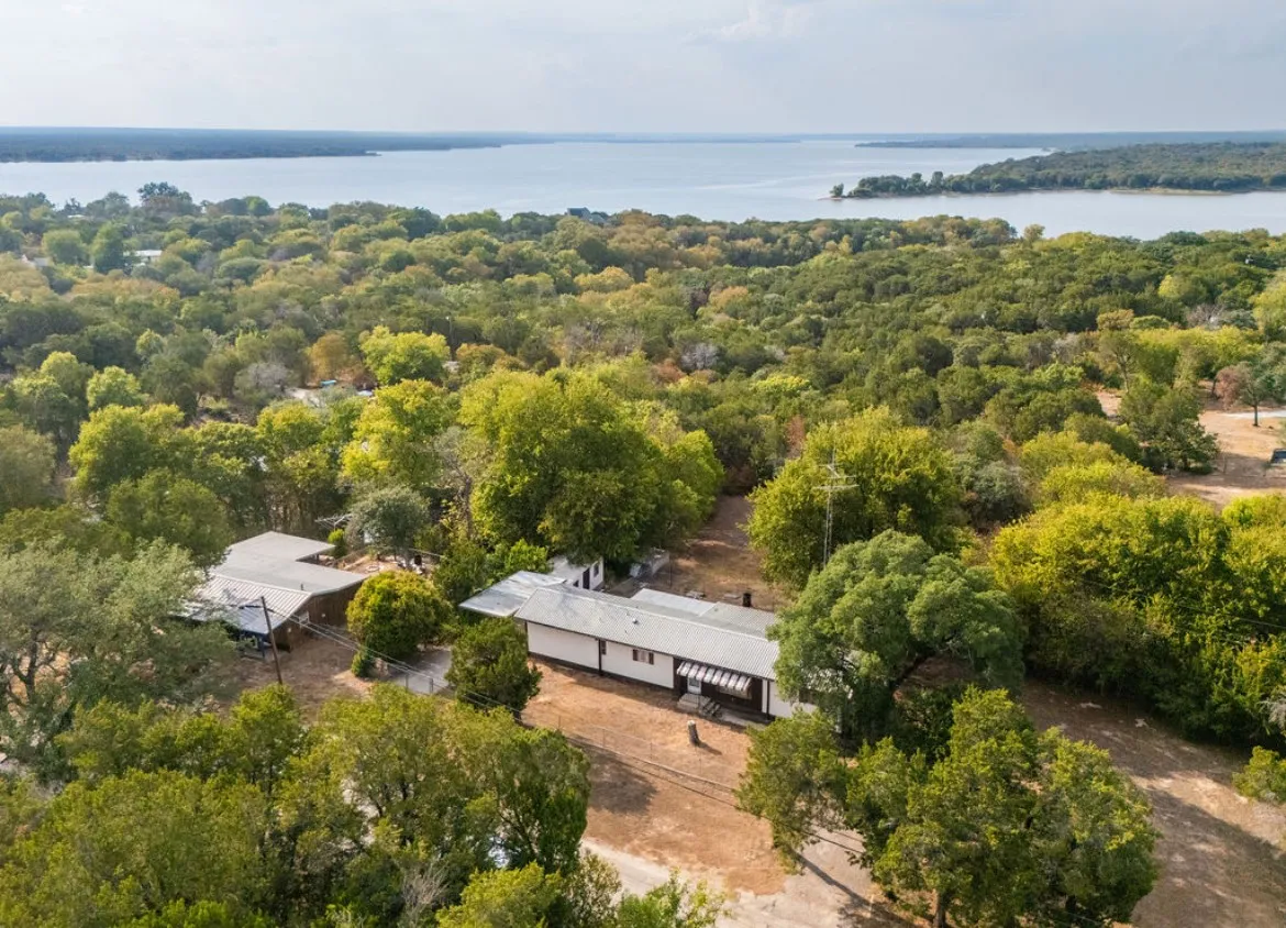 Expansive property featuring a single-story residence with a covered porch, surrounded by mature trees, with a prominent water body in the background