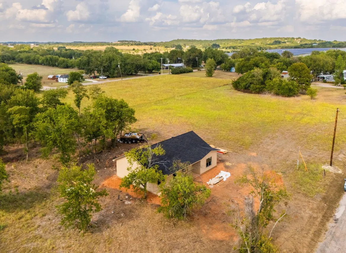 New construction home with a black shingle roof and light-colored siding, situated on a lot with mature trees and a paved road