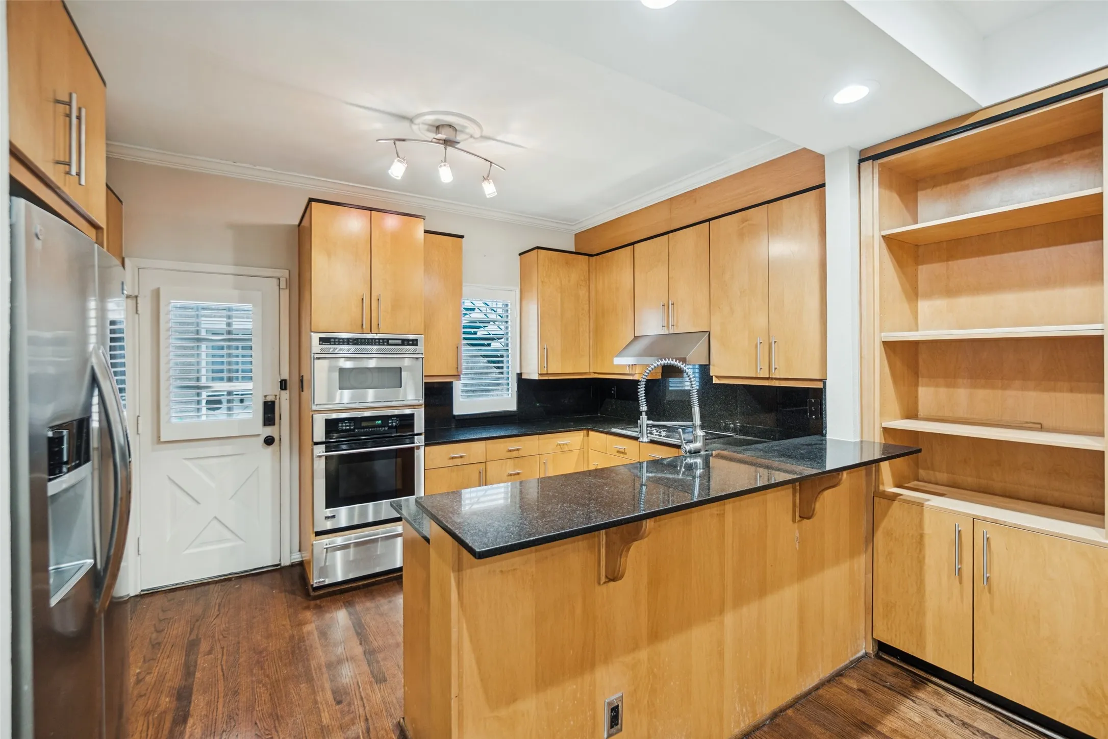 Kitchen featuring wood-finish cabinetry, stainless steel appliances, dark countertops, a built-in shelf unit, and hardwood flooring