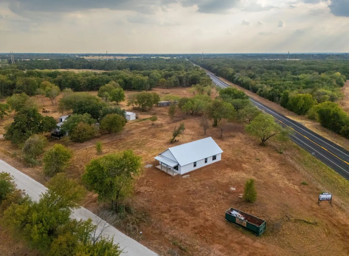 White exterior with a covered front porch, set on a sizable lot with mature trees, offering direct road frontage